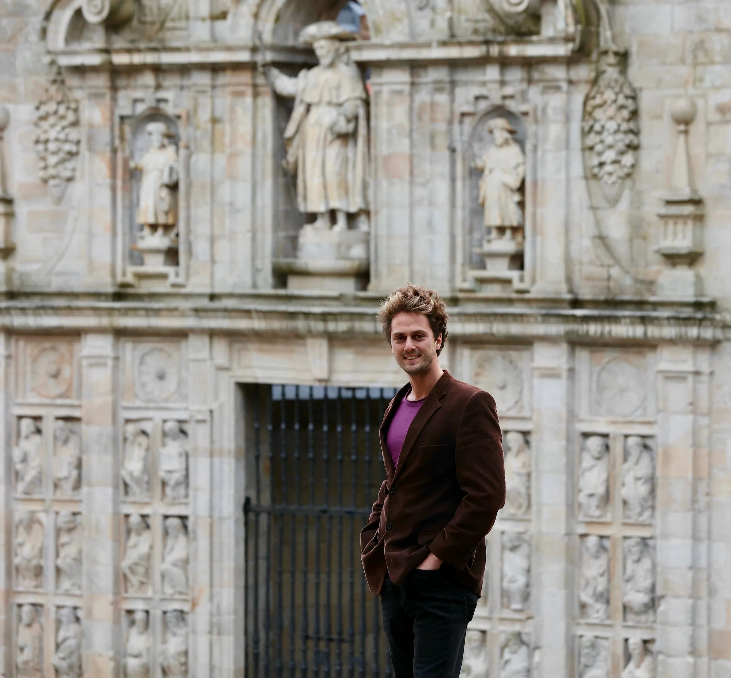 A young man in a brown blazer and purple shirt standing and smiling in front of a historic stone building with elaborate carvings and statues.