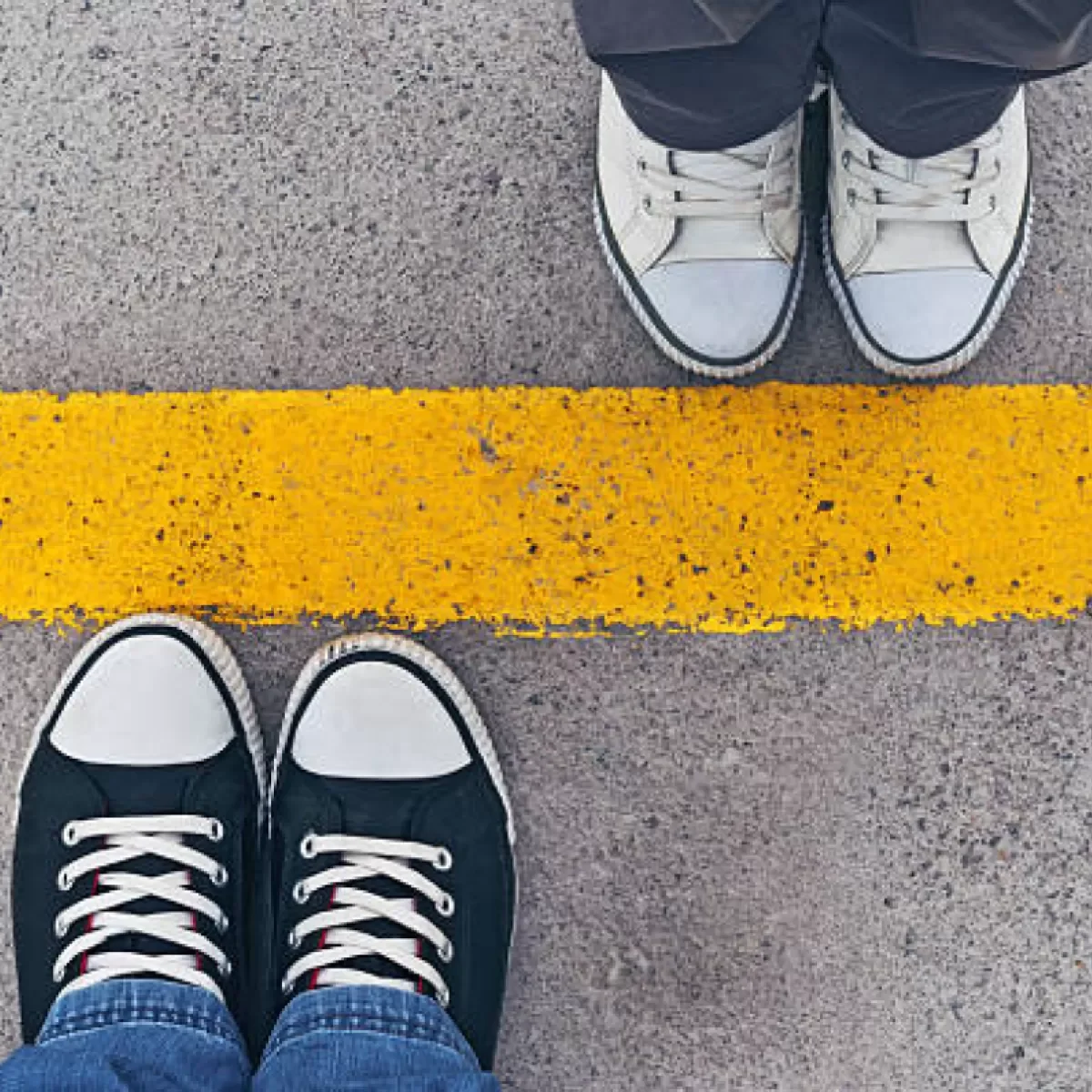 Two people in sneakers standing on a concrete sidewalk next to a yellow line.