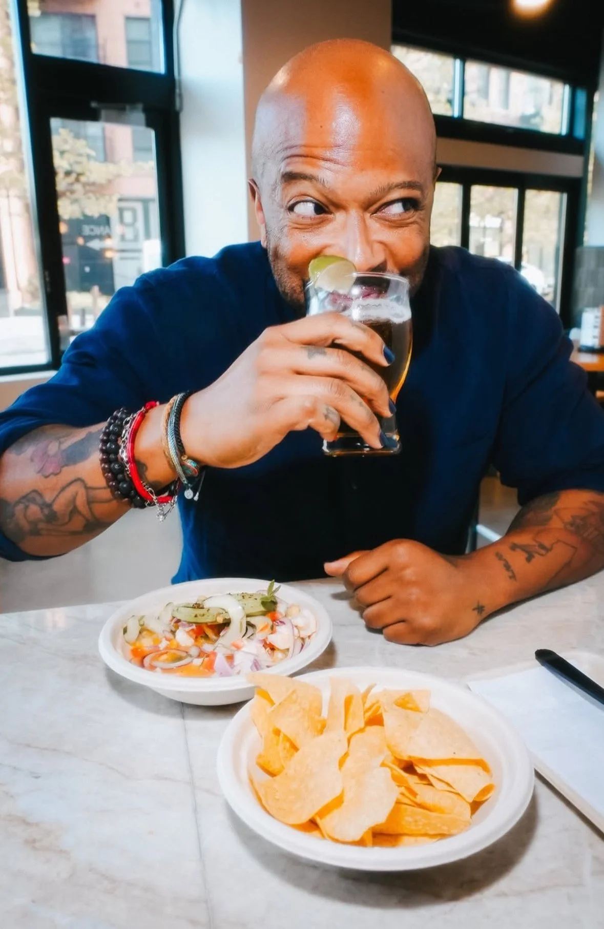 Kevin is wearing a blue oxford shirt and colorful bracelets while smiliing as he drinks a beer