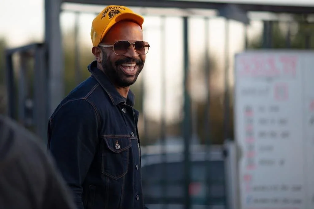 Kevin smiling at the camera wearing a yellow hat, denim jacket, and sunglasses.