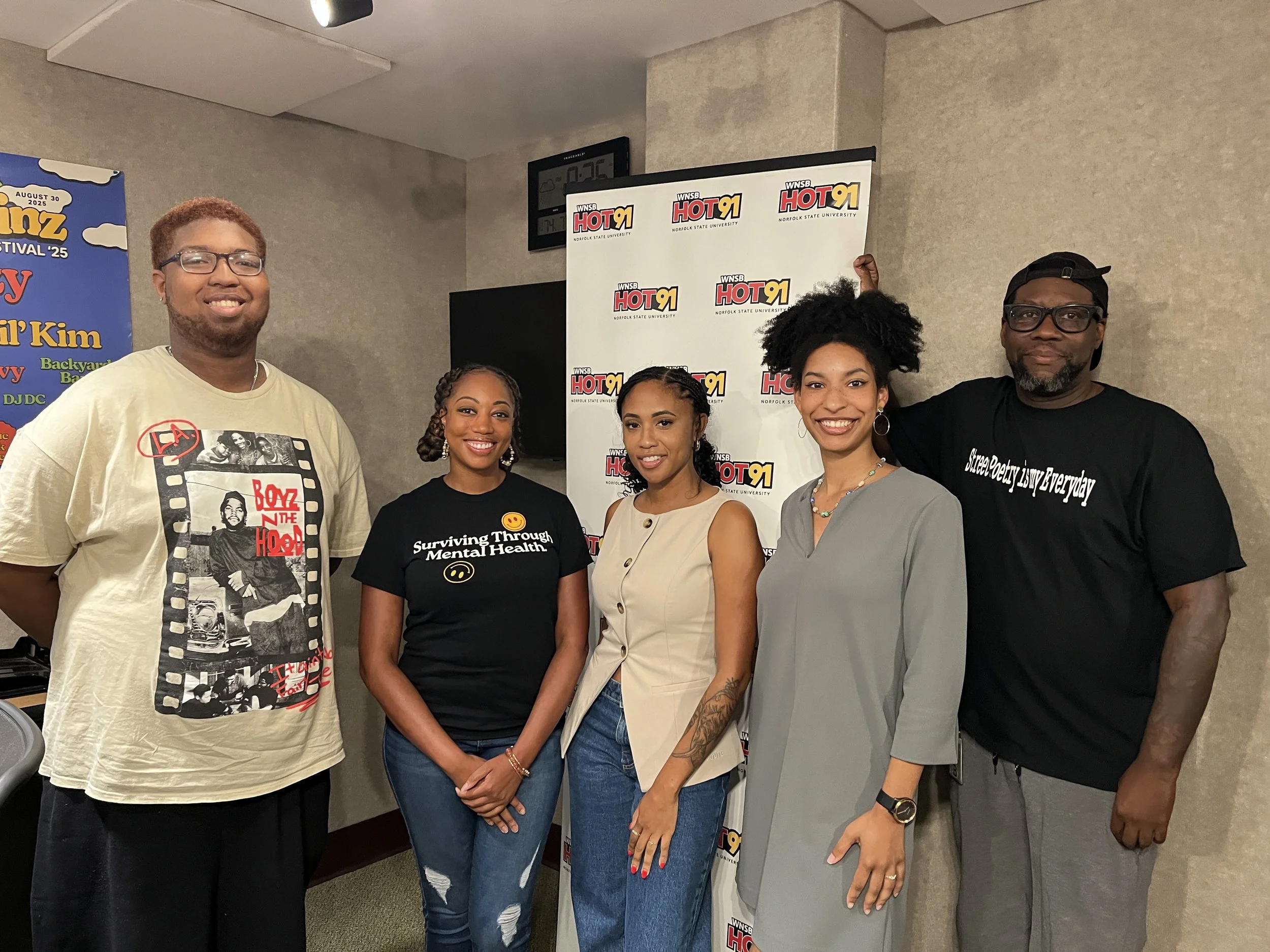 Group of five people standing in front of a backdrop with "WNSB Hot 91" logo, smiling at the camera, in an indoor setting with beige walls and a clock on the wall.