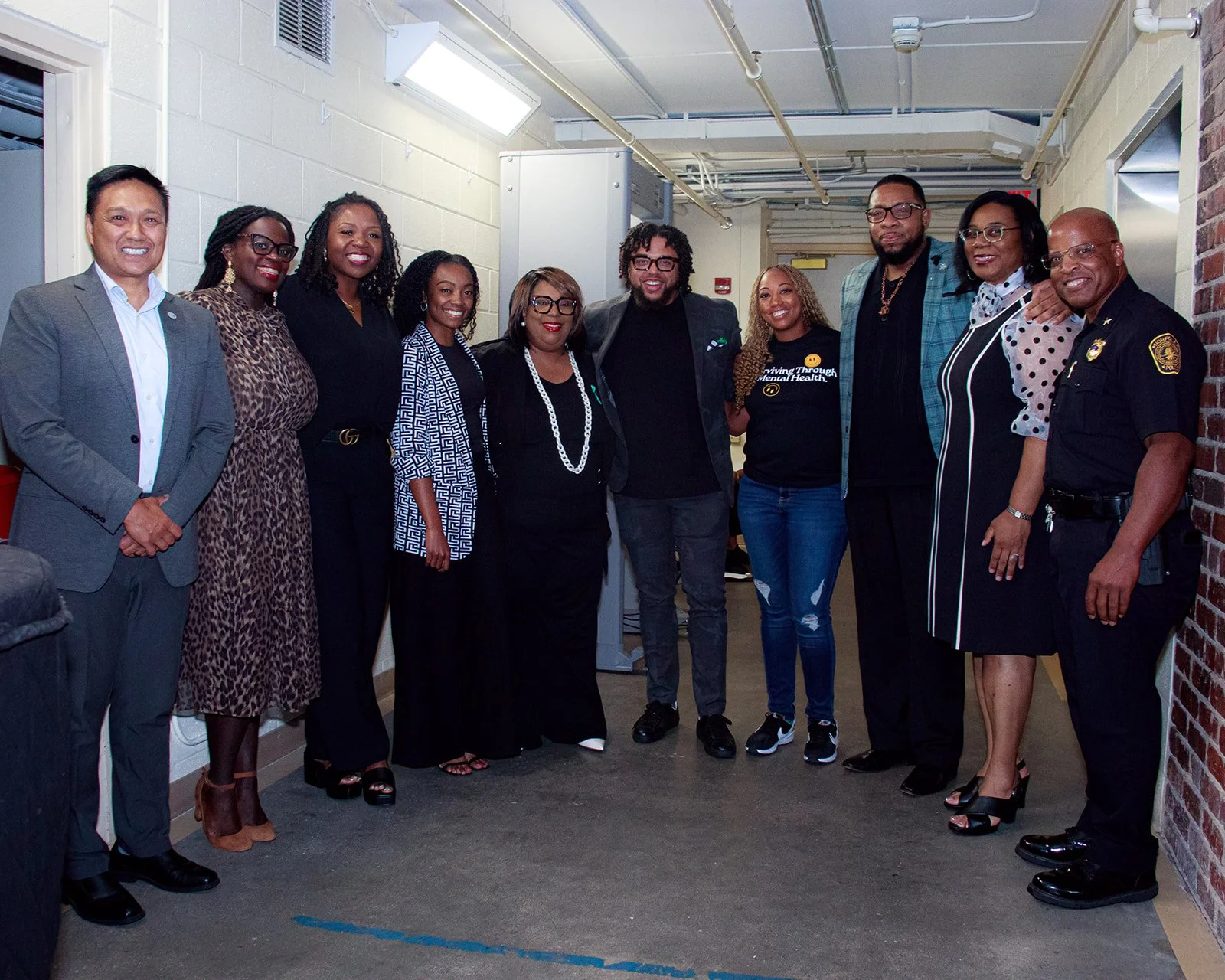 A diverse group of eleven people, including men and women, standing in a line in an indoor setting, smiling and posing for the photo. The group is dressed in business casual attire, with one person in a police uniform and another in a t-shirt with a message about mental health.