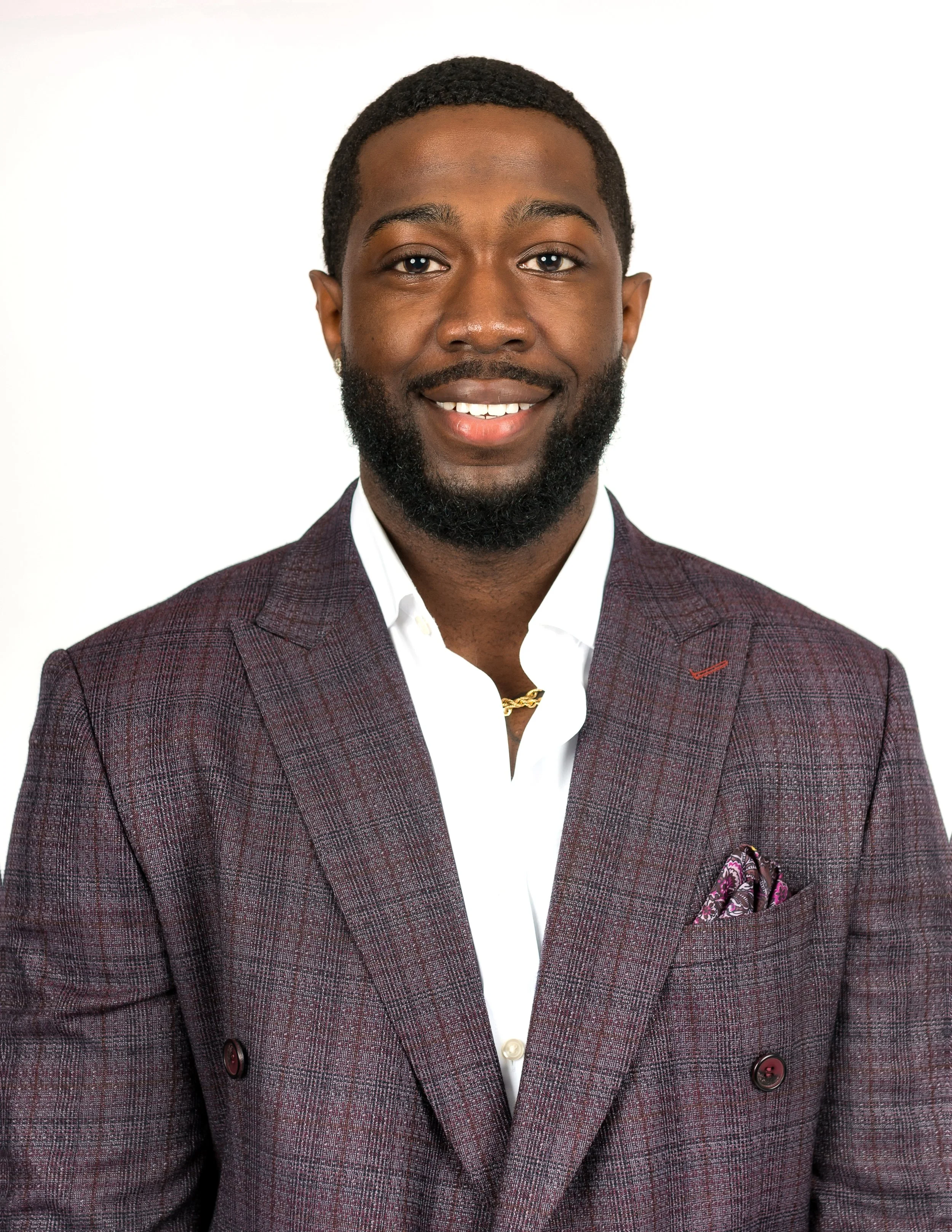 A professional man with dark skin, black hair, a beard, and a mustache, wearing a burgundy plaid blazer, white shirt, and gold necklace, standing against a white background.