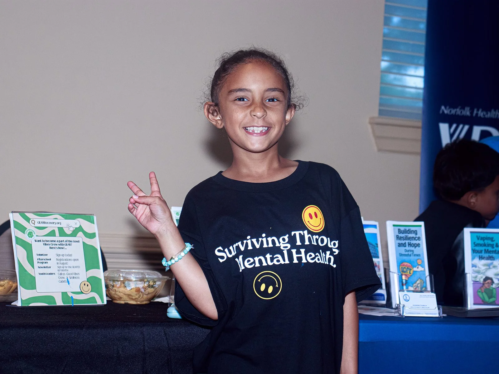 A smiling young girl with curly hair wearing a black T-shirt that reads 'Surviving Through Mental Health' with a smiley face graphic, making a peace sign with her right hand, standing in front of informational booths at an event.