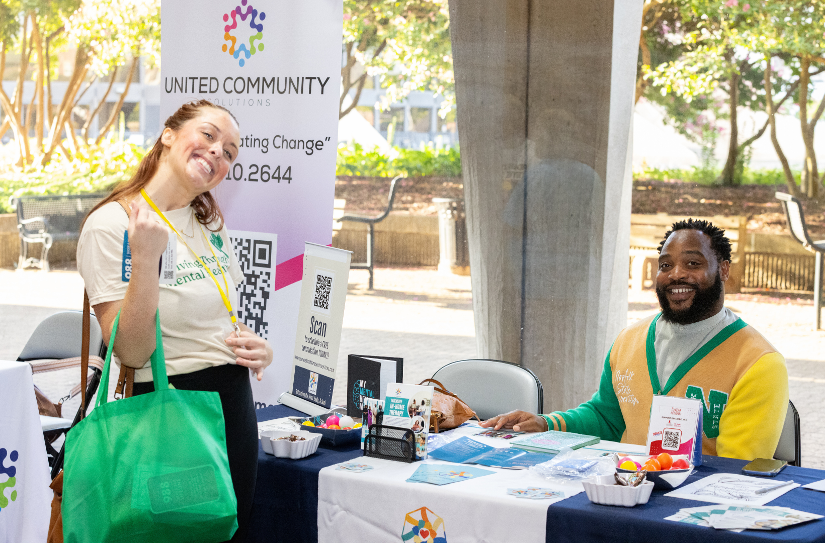 A woman with brown hair smiling and making a fist gesture at an outreach booth, and a smiling man with a beard sitting behind the table, at an outdoor event promoting United Community Solutions with informational brochures, QR codes, and a sign.