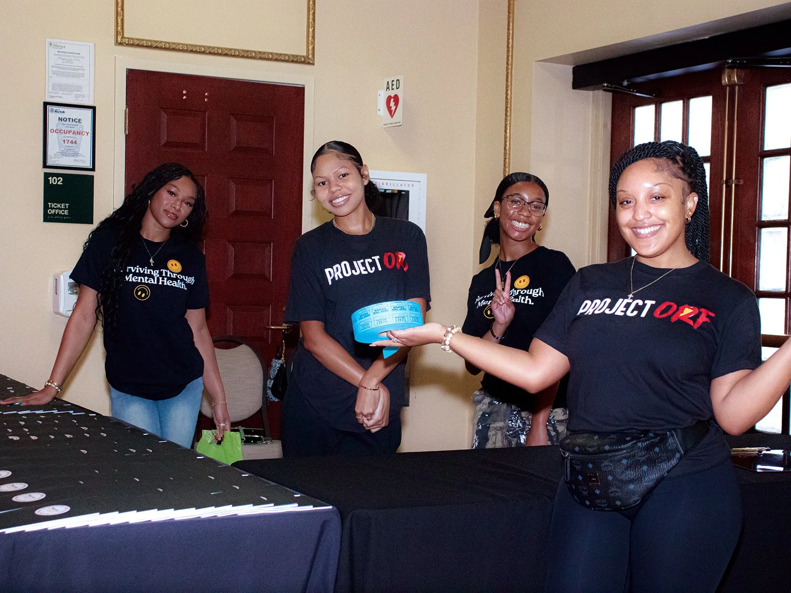 Four women standing together at an indoor event, smiling at the camera, wearing black t-shirts with health-related slogans, with a table in front of them and a wooden door and window in the background.