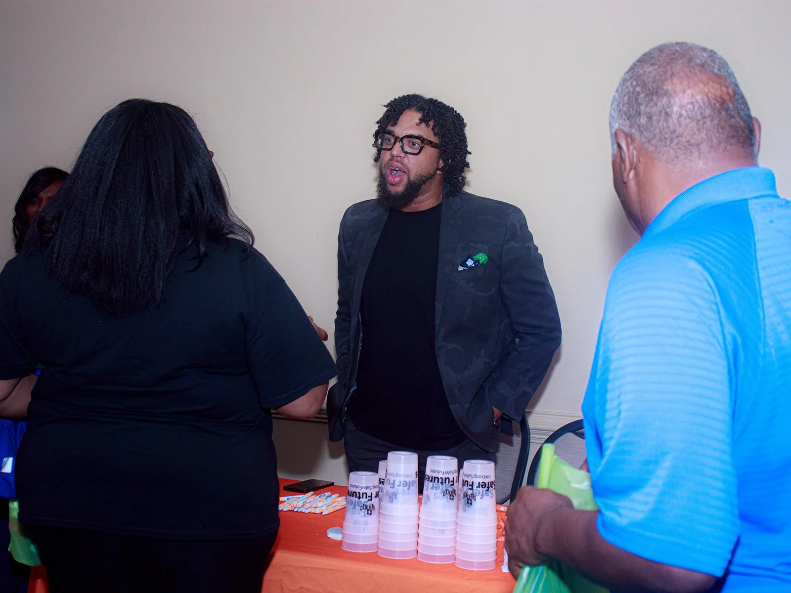 A man with glasses and a beard, in a dark blazer, is talking to two people at a table with orange cloth, which has cups, bottles, and other items on it. The woman with black hair and wearing a black top is facing him, and an older man with gray hair, in a blue shirt, is holding a green bag.