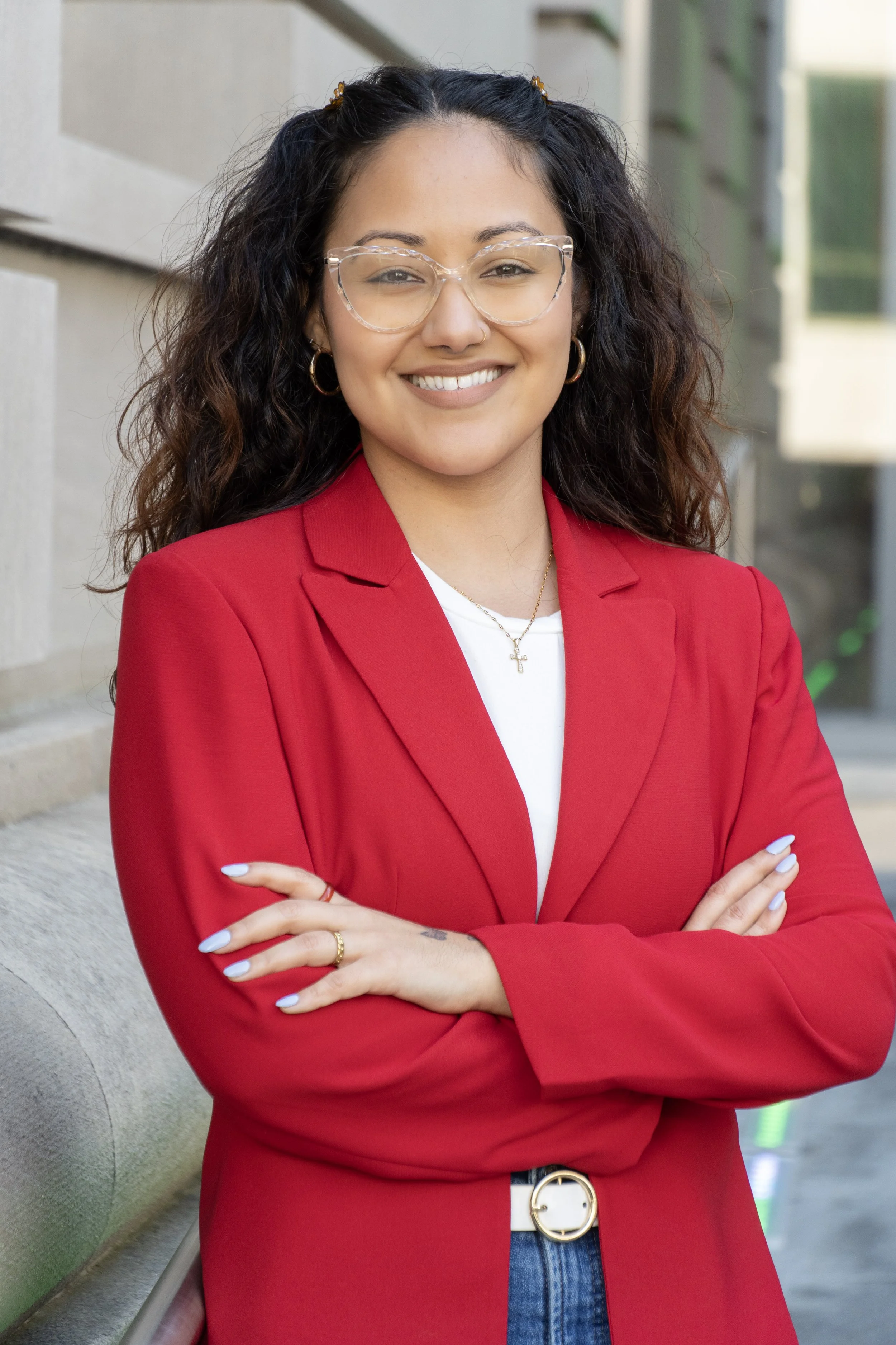 A woman with dark curly hair wearing glasses, a red blazer, a white shirt, and blue jeans, standing outdoors with arms crossed and smiling.