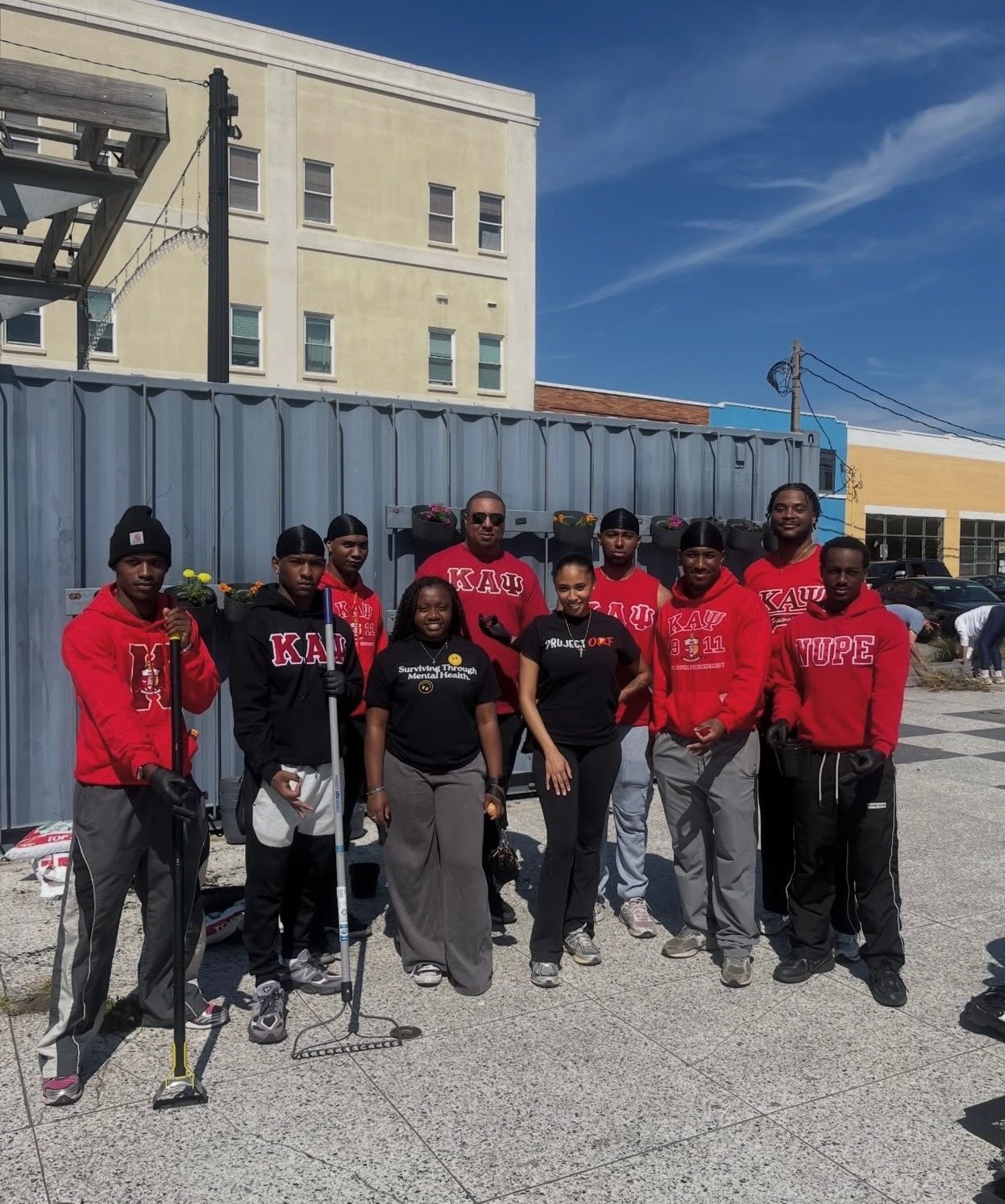 Group of people standing outdoors in front of a blue fence, some wearing red hoodies with Greek organizations names, holding trash pickers, with a building and parked cars in the background.
