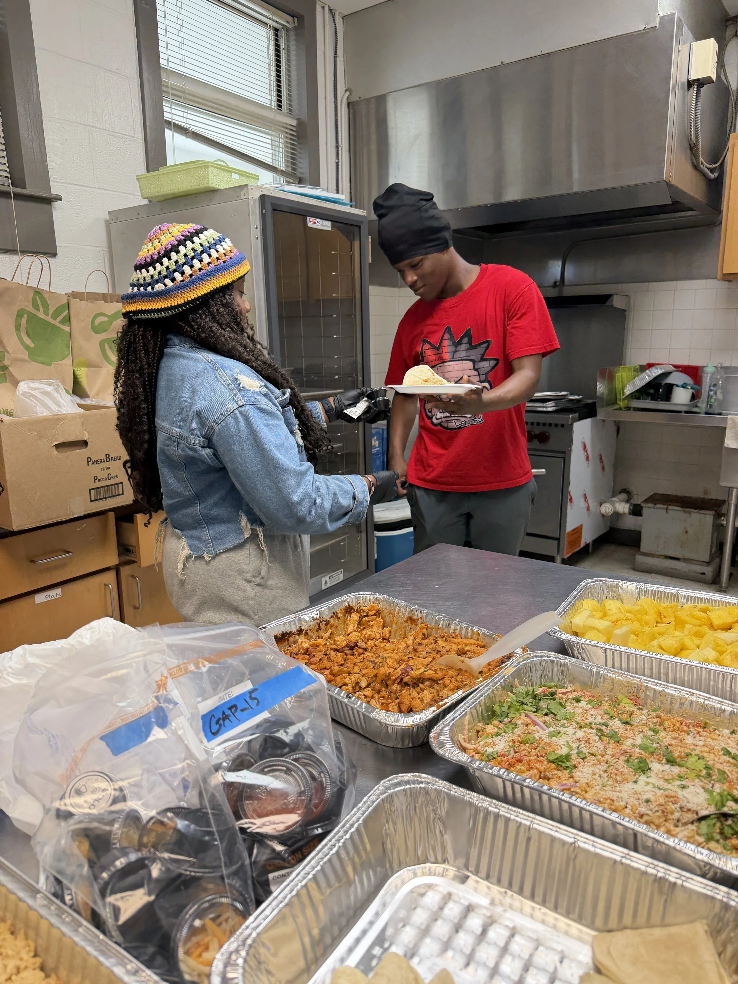 A person wearing a multicolored crocheted hat and denim jacket receives food from another person in a kitchen setting, with trays of various dishes, including beans, rice, and vegetables, on a table in the foreground.