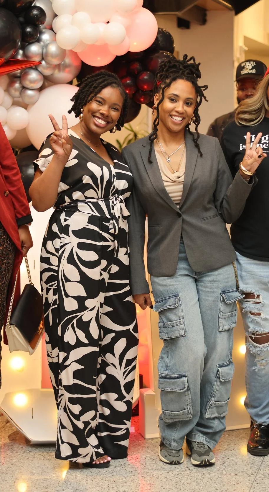Two women standing together, smiling and making peace signs. The woman on the left wears a black and white patterned jumpsuit, and the woman on the right wears a gray blazer with cargo jeans. They are at an indoor event with black, white, pink, and red balloons in the background.