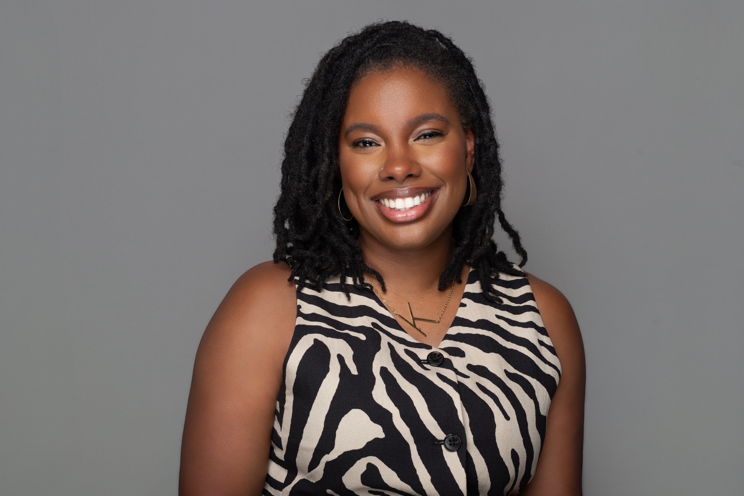 A woman with dark, curly hair styled in dreadlocks, smiling and wearing a zebra print sleeveless top and gold jewelry, against a plain grey background.