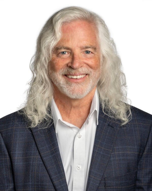 A smiling older man with long white hair and a beard, wearing a white shirt and dark blazer, against a white background.