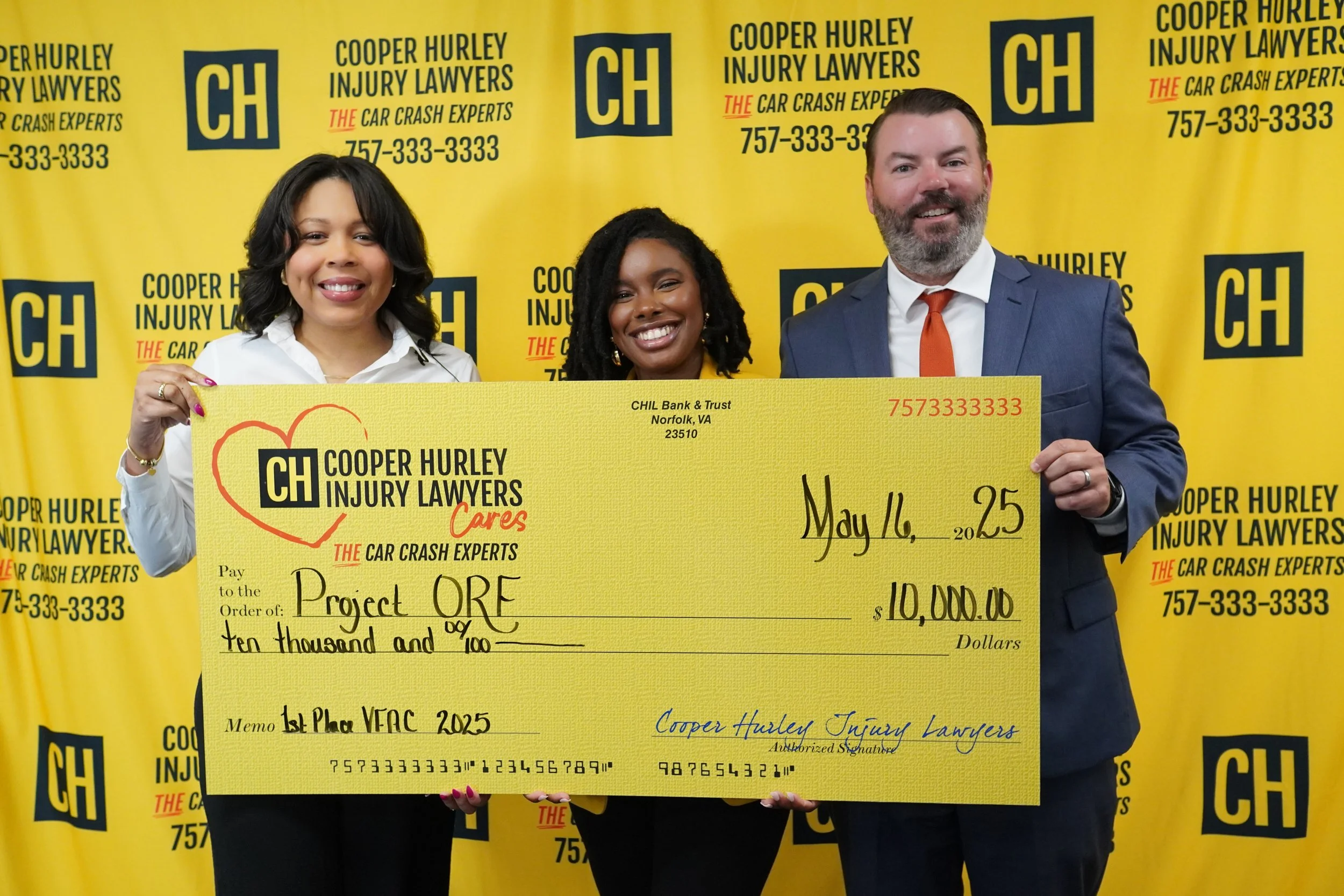 Three people holding large yellow check in front of yellow background with black text. The woman on left is smiling, wearing a white top and gold bracelet. The woman in middle is smiling, wearing earrings and a yellow outfit. The man on right is smiling, in a suit with a white shirt and orange tie.