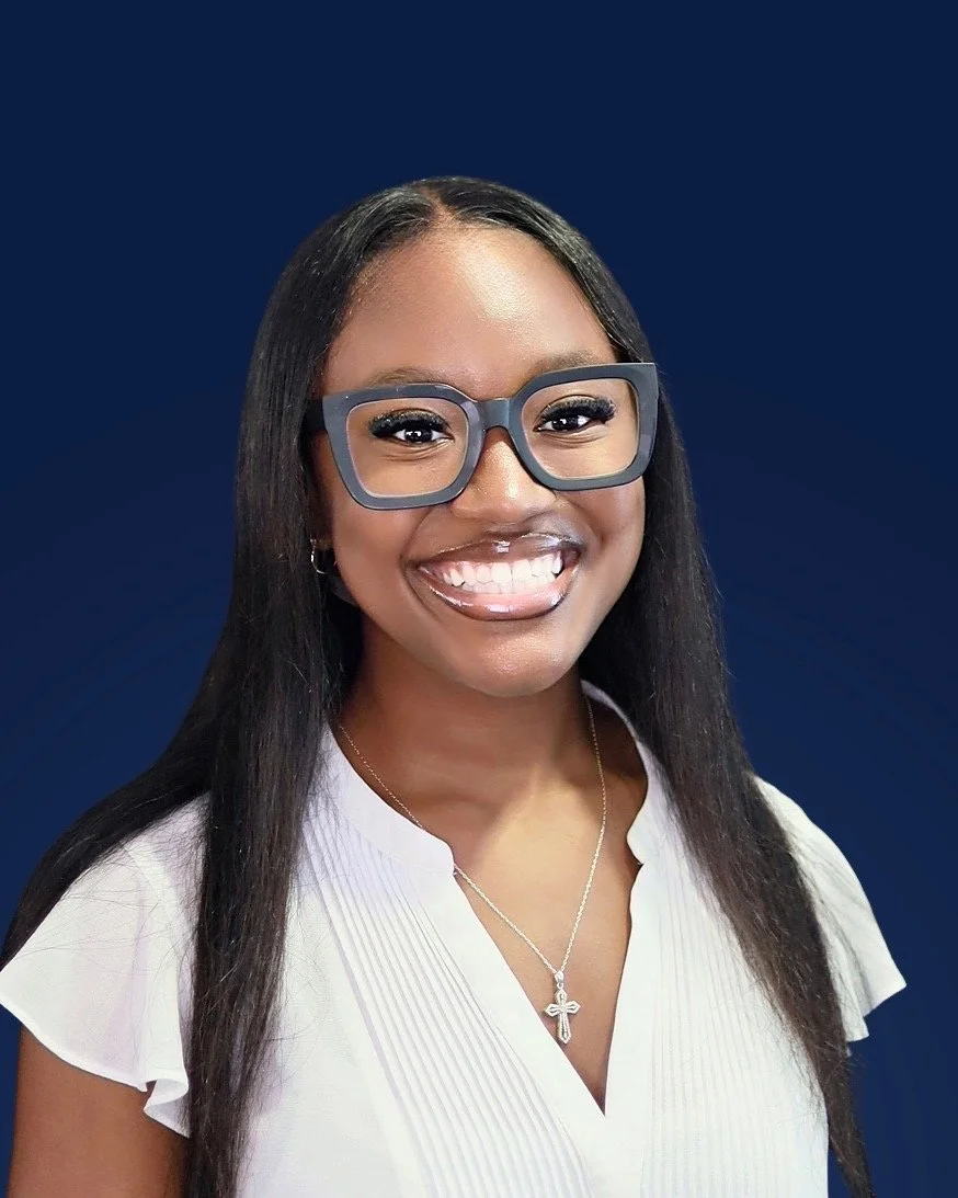 A young woman with long dark hair, wearing glasses, a white blouse, and a necklace with a cross pendant, smiling against a dark blue background.