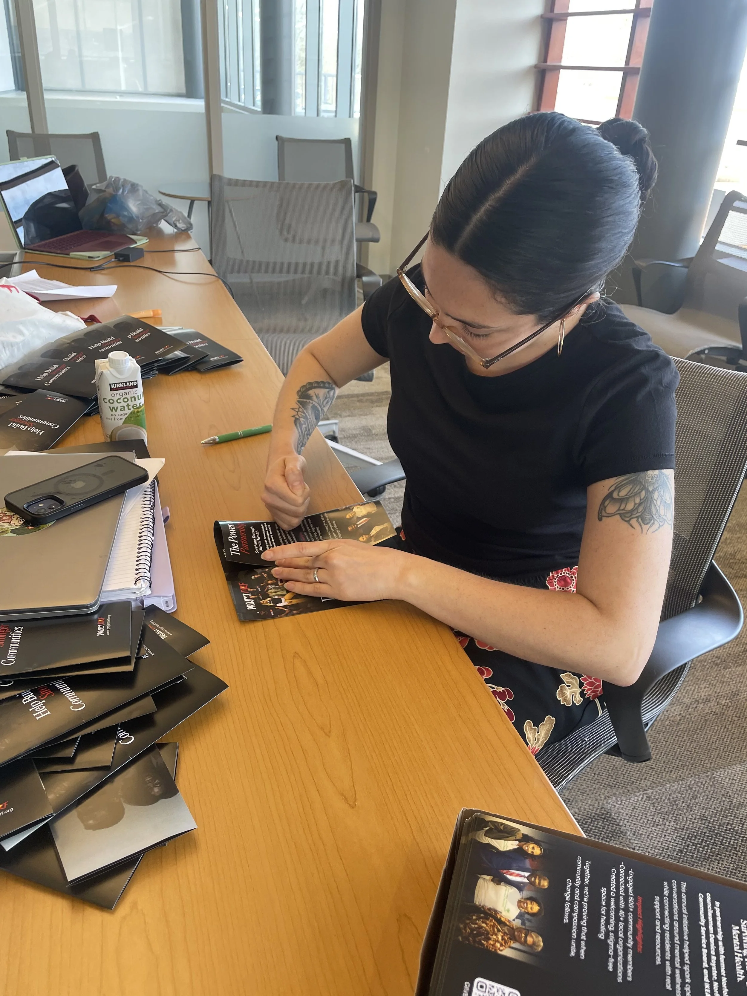 A woman with dark hair in a bun, glasses, and tattoos on her arm, sitting at a wooden table, looking at and flipping through a magazine in an office or conference room. The table has multiple black pamphlets, a phone, a notebook, a pen, and a bottle of coconut water on it. There are laptops and chairs around the room, and large windows with natural light.
