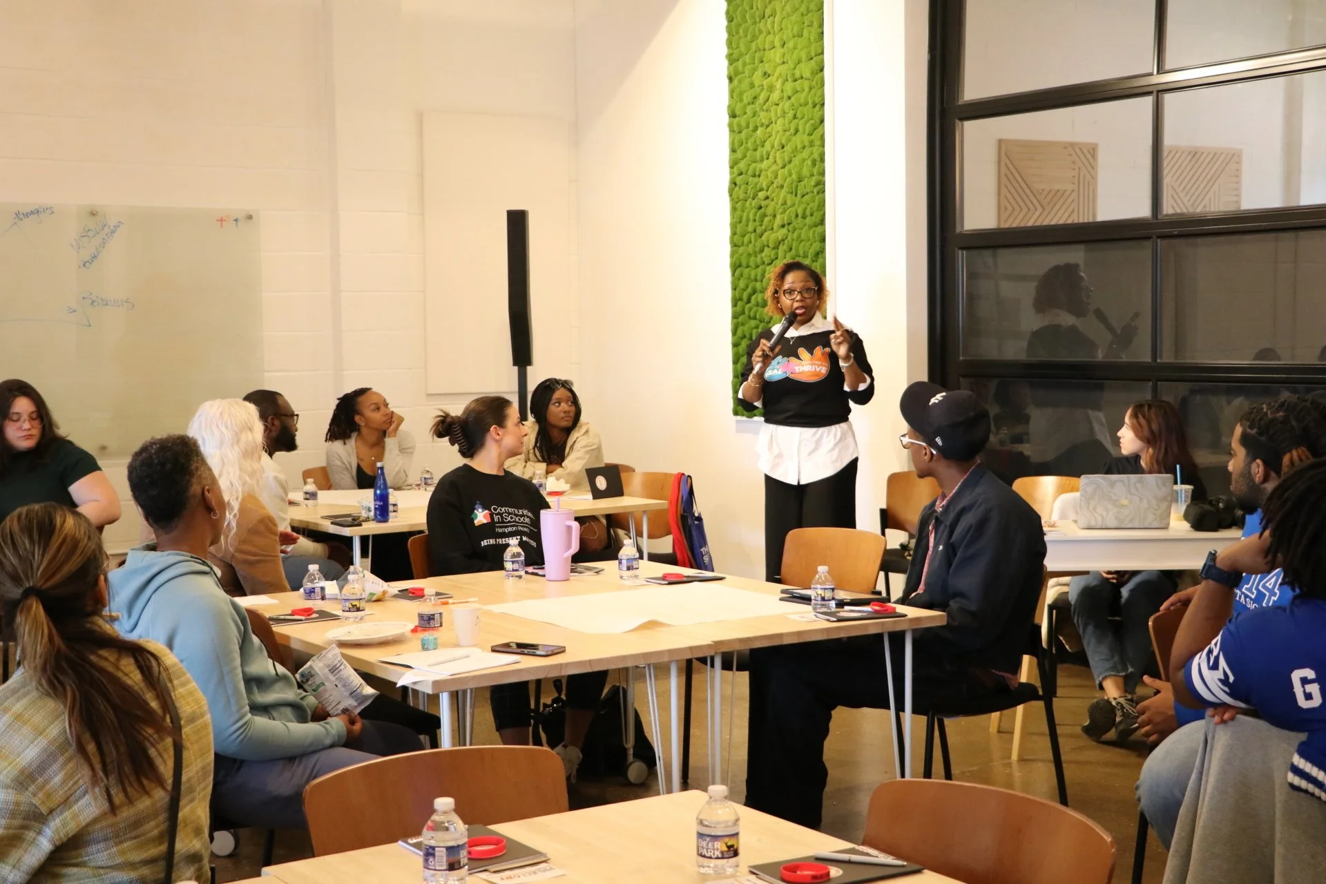 A woman is speaking into a microphone during a presentation in a well-lit room with attendees seated at tables, some with water bottles, notebooks, and laptops, and a decorative green moss wall in the background.