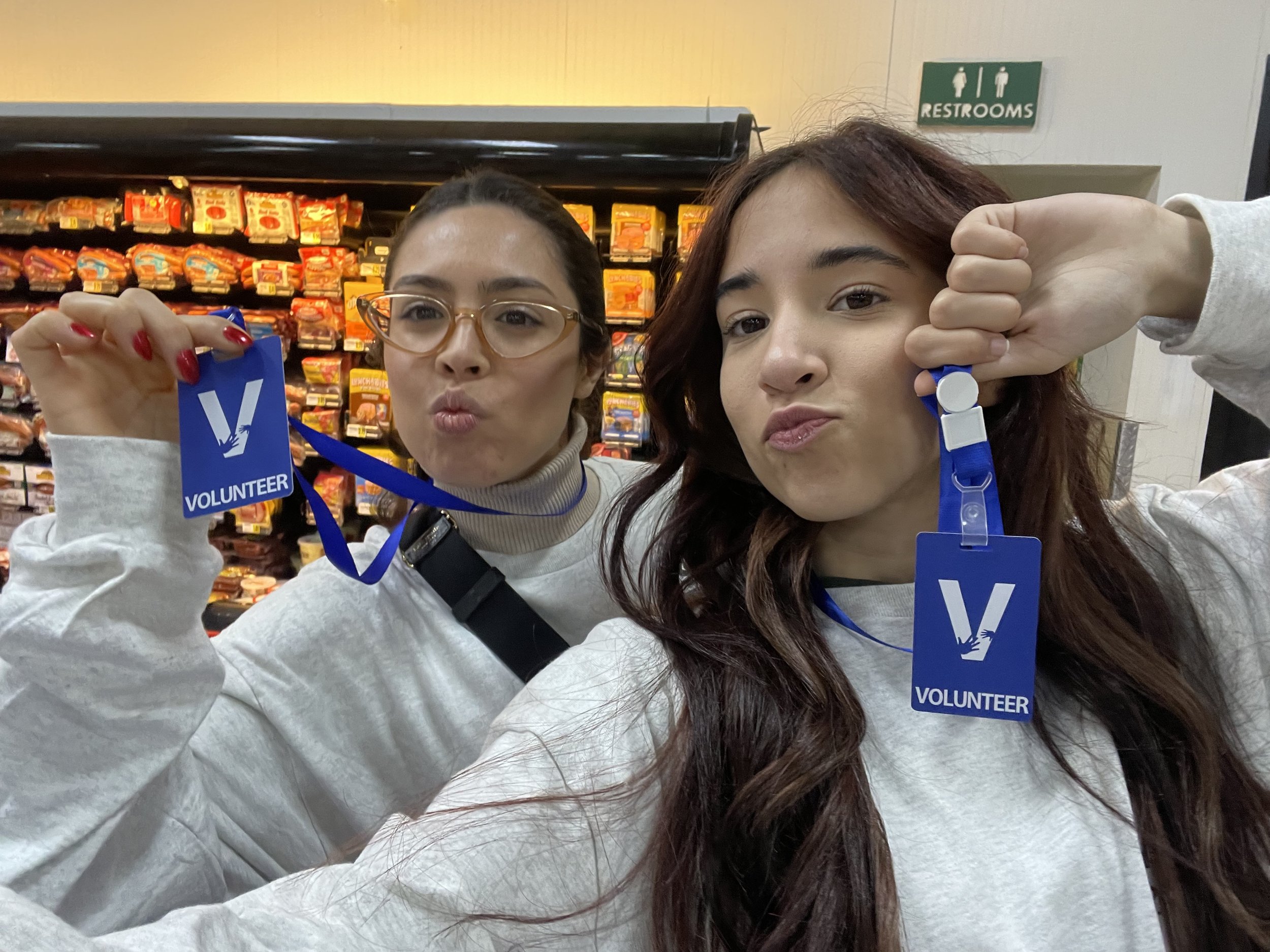 Two women taking a selfie in a grocery store, holding volunteer badges. Shelf with snacks in the background, and a restroom sign in the upper right.