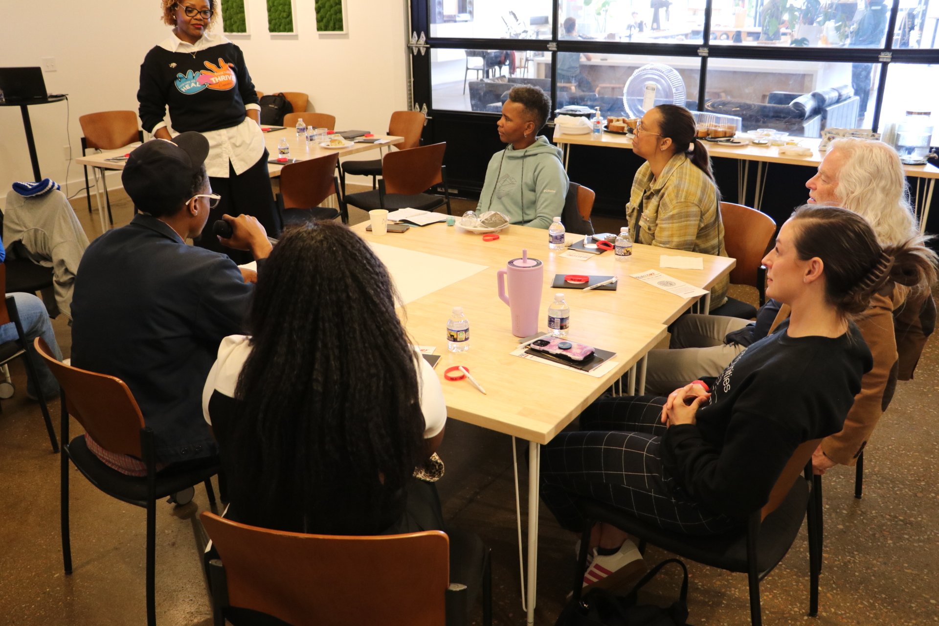 A woman standing and speaking to a group of people sitting at a large table in a conference room. The group appears engaged, with various items such as water bottles, snacks, and notebooks on the table. Large windows are visible in the background, and the room has simple decor with green wall art.