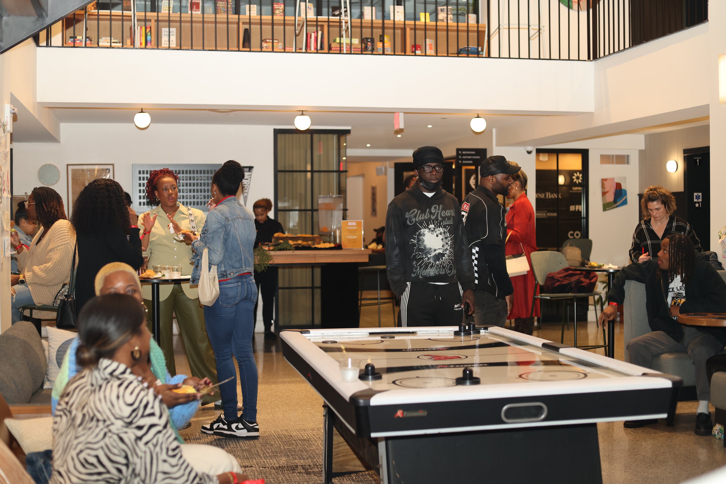 People gathered in a modern indoor space, some standing and talking, others sitting and eating, with a large air hockey table in the foreground and a bookshelf on the upper level in the background.