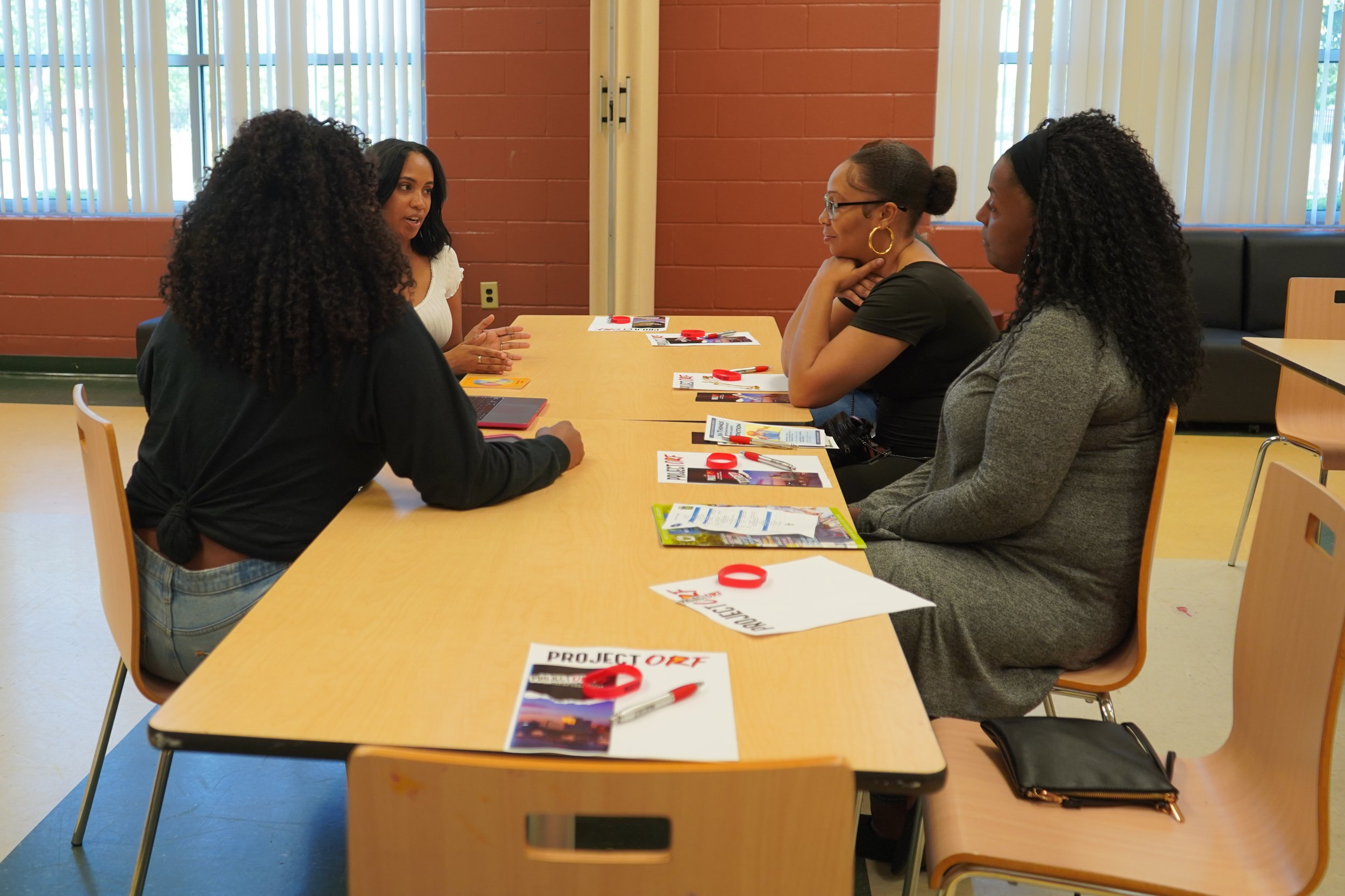 Four women are sitting at a table engaged in a discussion, with brochures, pens, and red wristbands on the table inside a room with large windows and beige curtains.