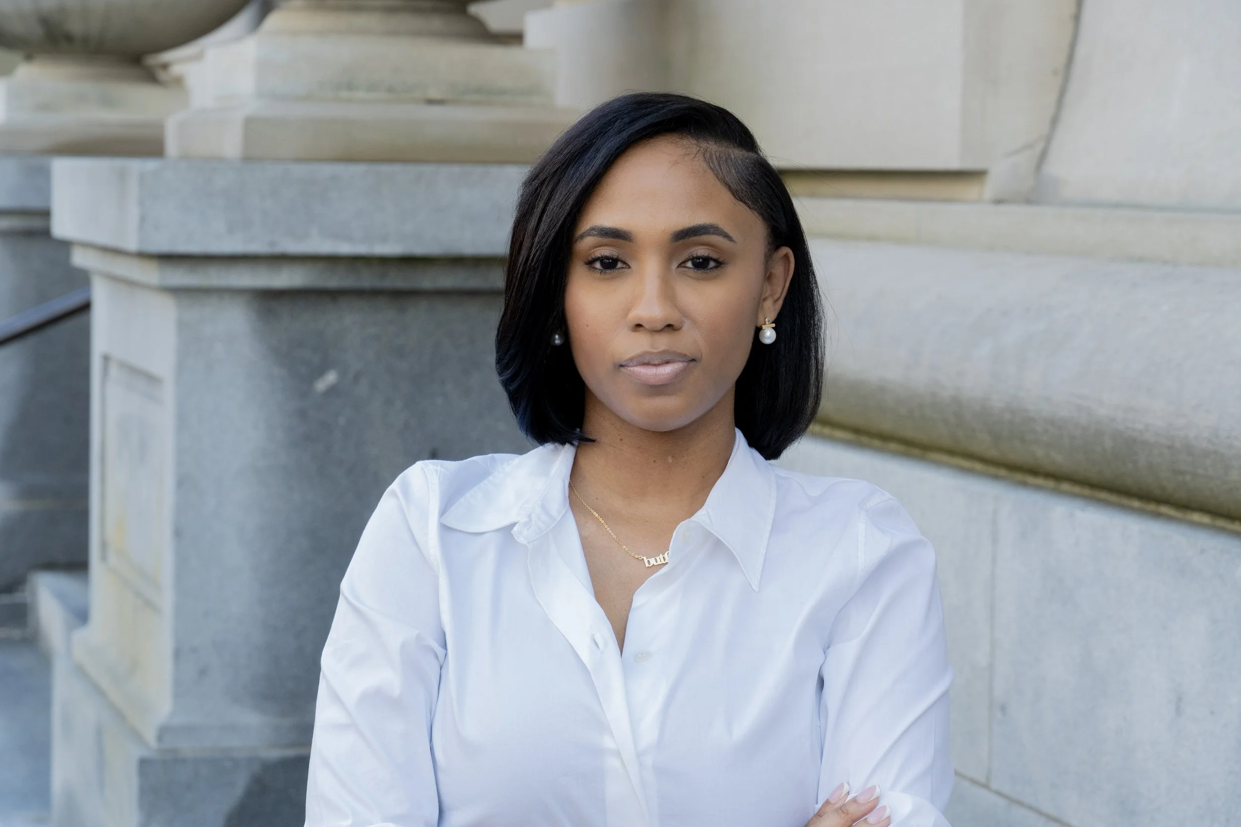 A woman with short black hair and pearl earrings poses outdoors, wearing a white button-up shirt, in front of a stone structure.