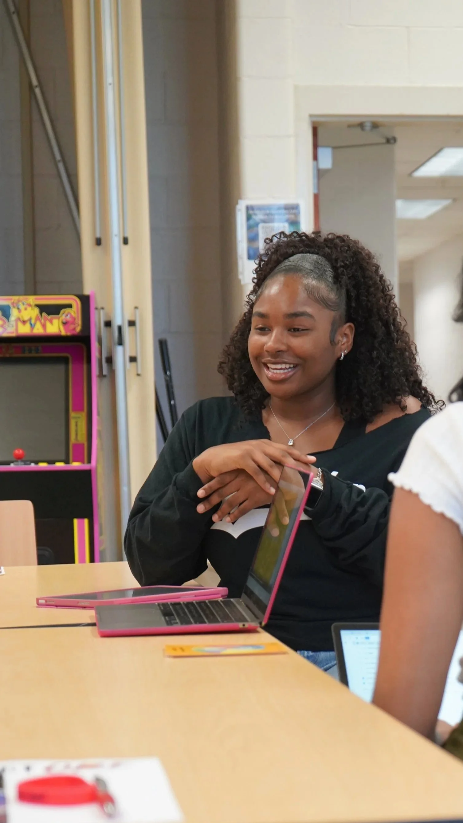 A young woman with curly hair and a gray headband, smiling and sitting at a table with a pink laptop, in a room with arcade machines and a beige wall.