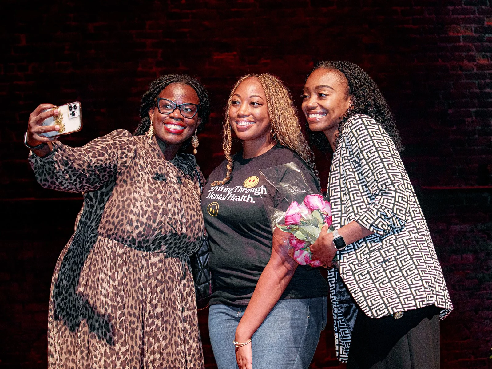 Three women standing together taking a selfie. The woman on the left is wearing glasses and a leopard print dress. The woman in the middle is holding a bouquet of pink flowers and wearing a black t-shirt with yellow and white text and symbols, smiling at the camera. The woman on the right is wearing a black and white geometric patterned shirt and holding the bouquet, also smiling.