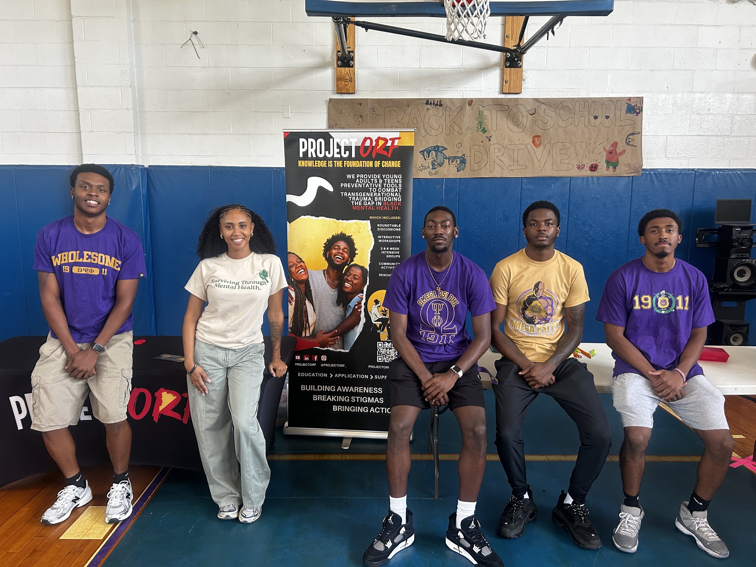 A group of five young people standing in front of a banner that reads 'Back to School Drive'. They are in a gymnasium with a basketball hoop in the background. Some are wearing purple shirts with Greek letters, and others are dressed casually.