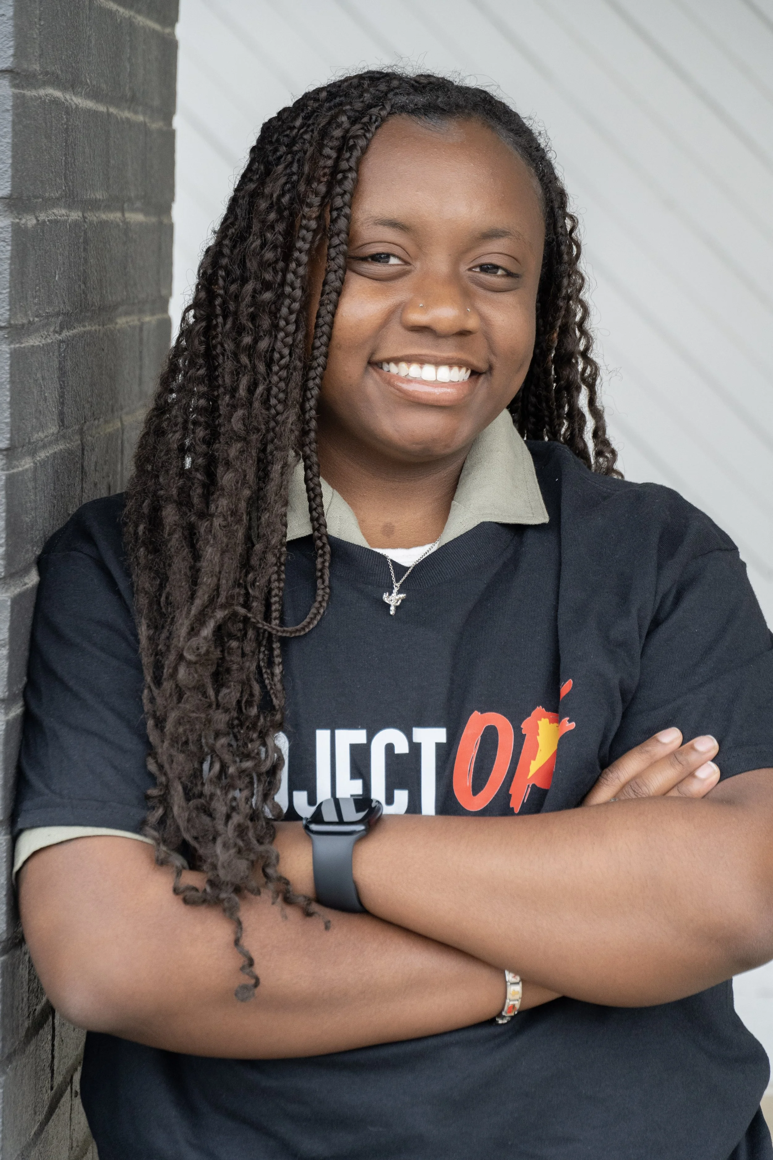 A smiling young woman with braided hair standing outdoors against a gray brick wall. She is wearing a dark t-shirt with a colorful logo and has her arms crossed.