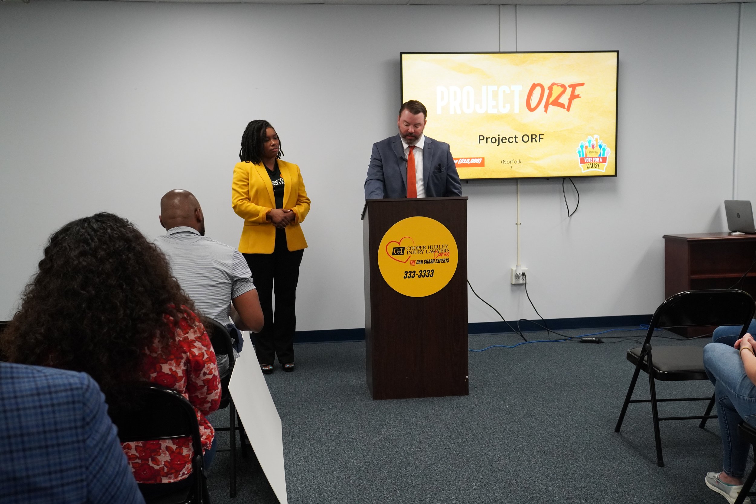 A man in a suit and a woman in a yellow blazer are giving a presentation in a conference room. The man is standing at a podium with a logo and phone number, while the woman stands beside him. Behind them, a large screen displays the words 'PROJECT ORF' and 'Vote for a Cause' with multiple hands raised graphic.