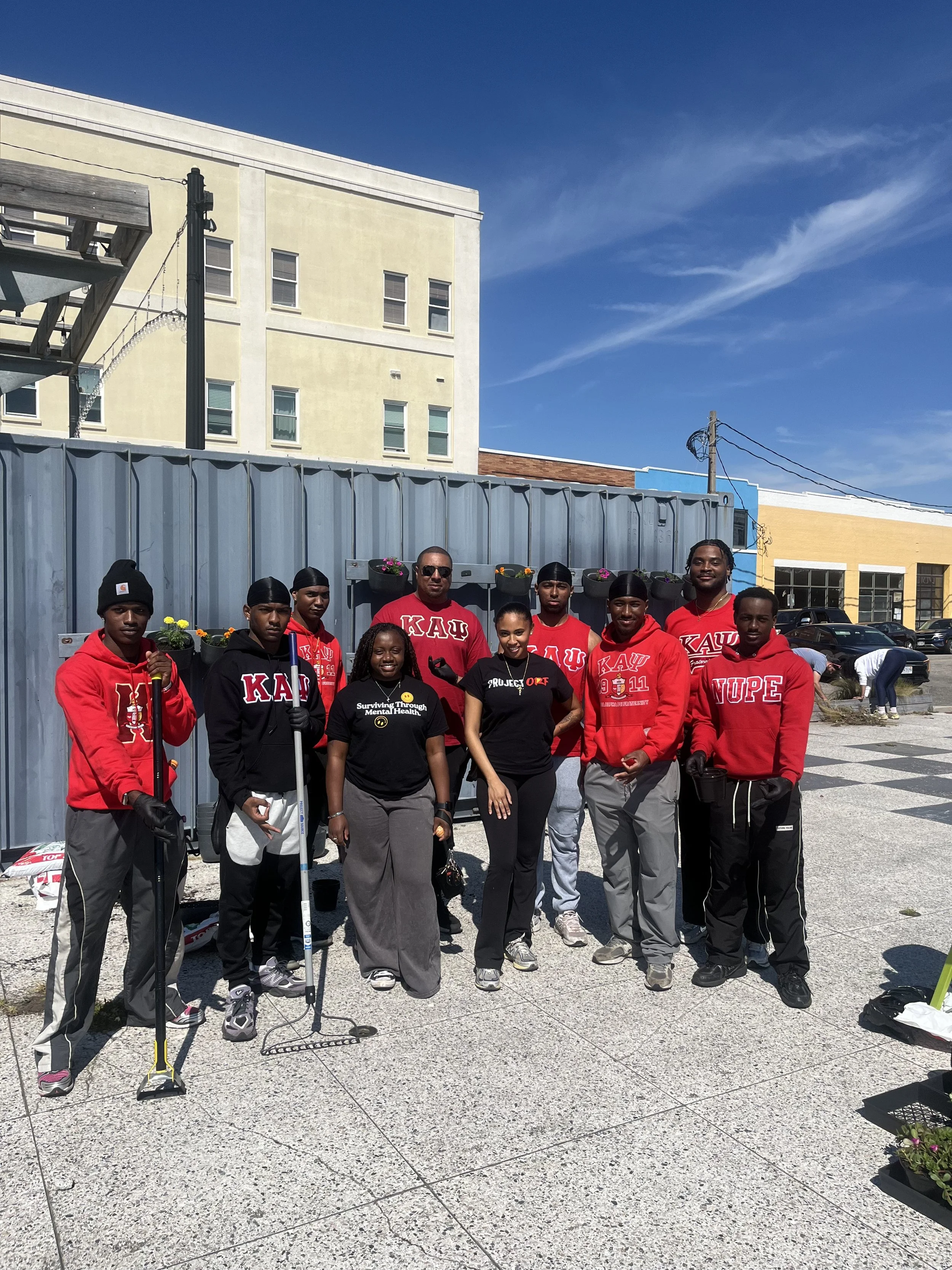Group of young adults wearing red and black shirts with Greek letters, standing outdoors in an urban area, some holding tools for planting or cleaning, in front of a gray fence and buildings under a blue sky.