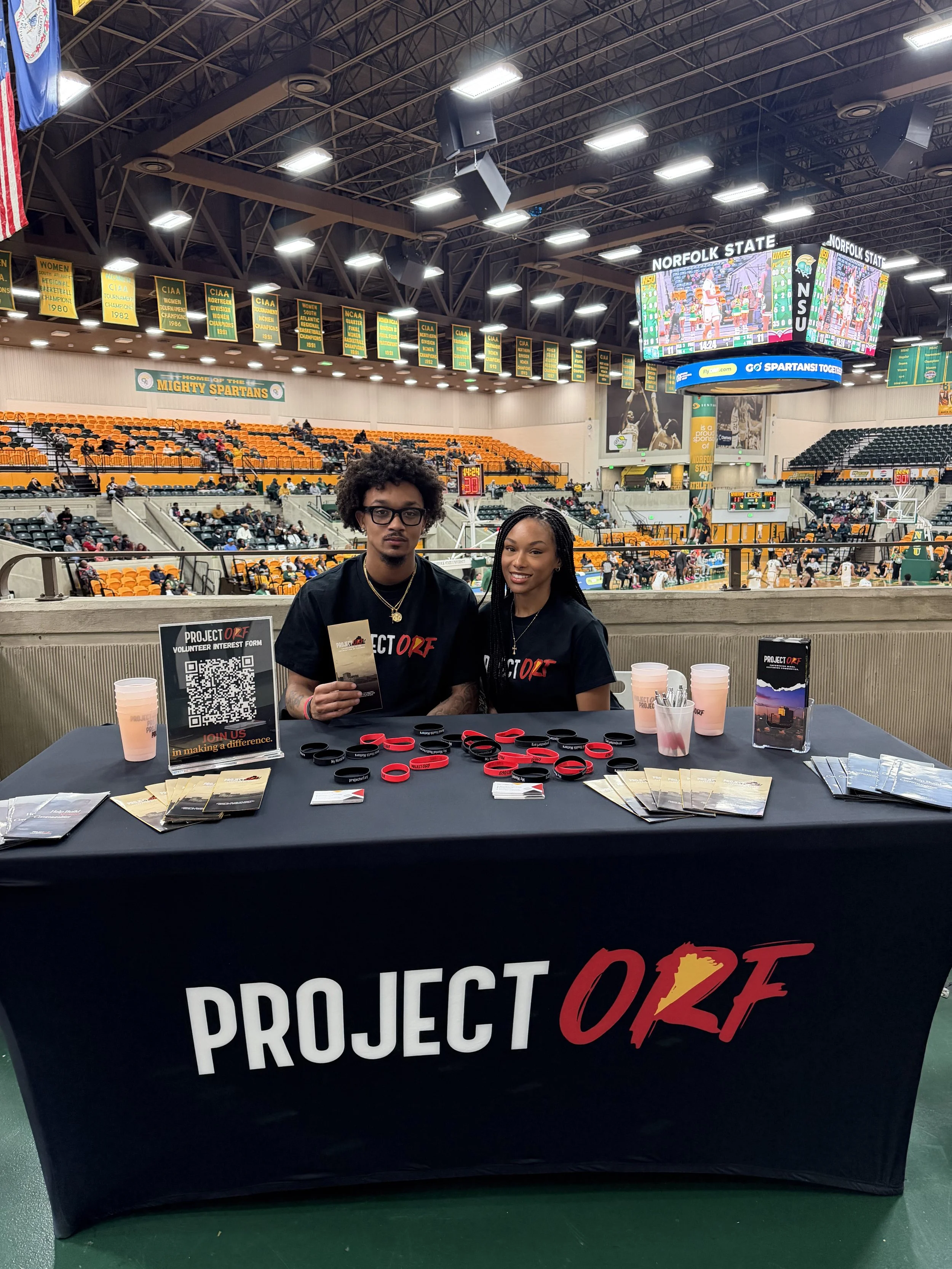 Two individuals sitting behind a table promoting Project OAK at an indoor sports arena, with banners, spectators, and a scoreboard in the background.