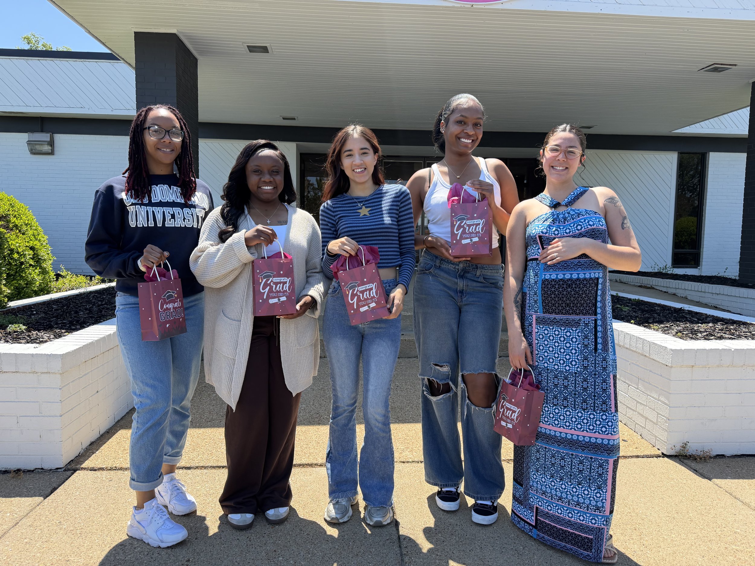 Six women standing outside in front of a building, holding pink gift bags and celebrating graduation. They are smiling and dressed casually, with some wearing glasses and necklaces.