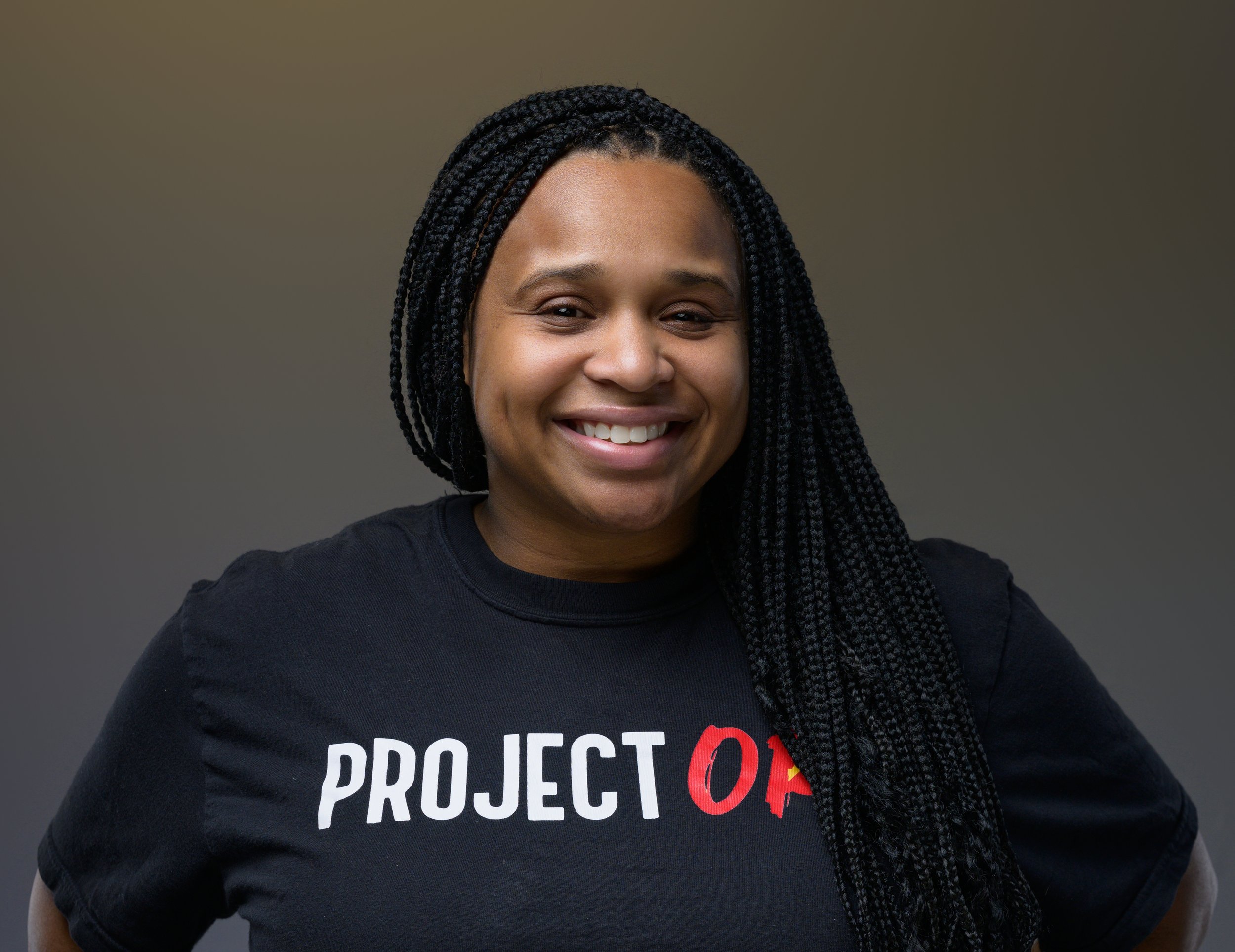 A woman with braided hair wearing a black t-shirt that says 'PROTEST' in white and red letters, smiling against a plain background.