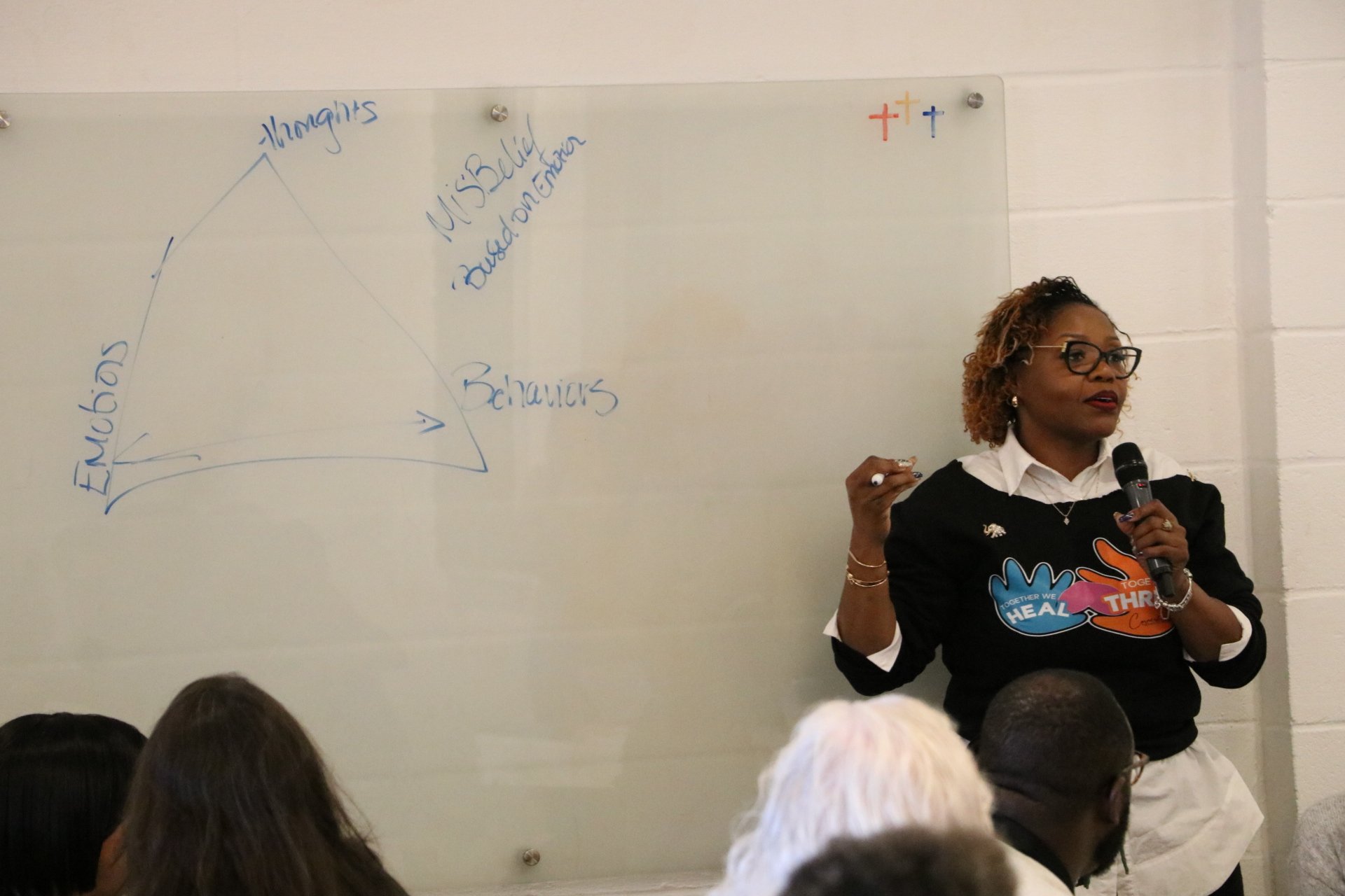 A woman with curly hair, glasses, and earrings is giving a presentation in front of a whiteboard, holding a microphone and a marker. The whiteboard has handwritten notes and colored symbols. The audience is visible from the back of their heads.
