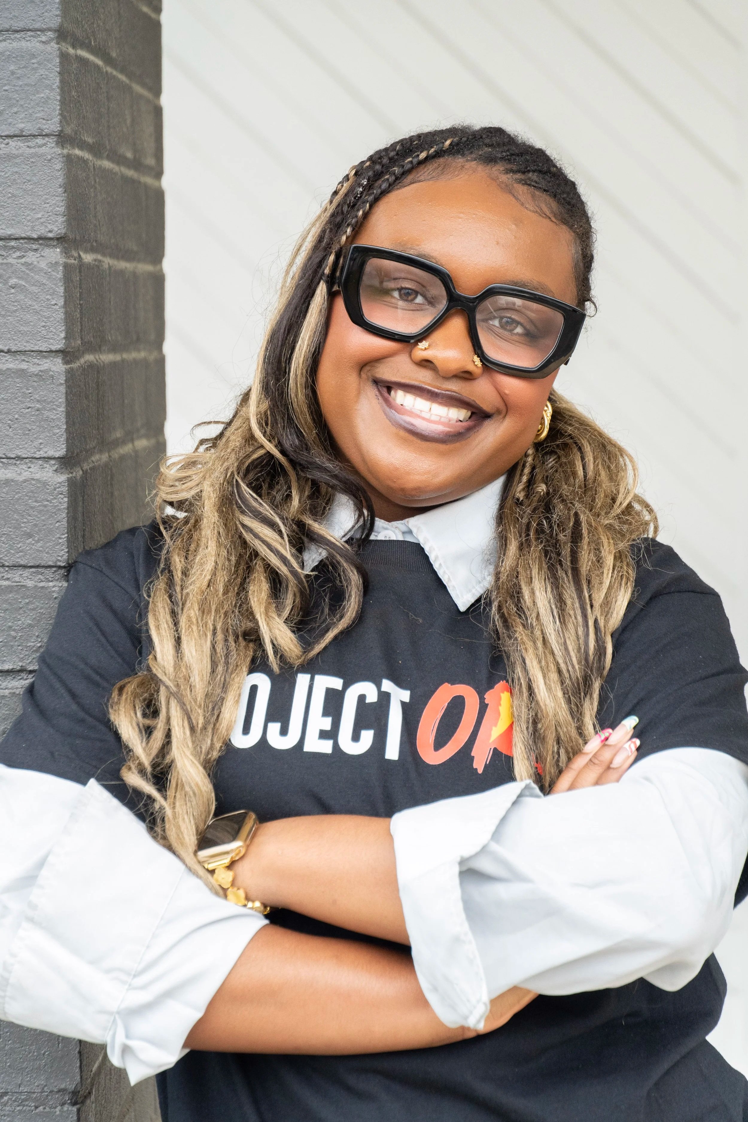 A woman smiling with crossed arms, wearing large black glasses, a necklace, earrings, a white collared shirt under a black T-shirt, standing next to a gray brick wall and a white wall with diagonal shadows.