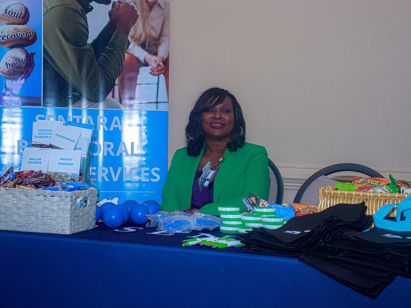 A woman sitting at a table with mental health awareness materials, snacks, and promotional items at a mental health services event.