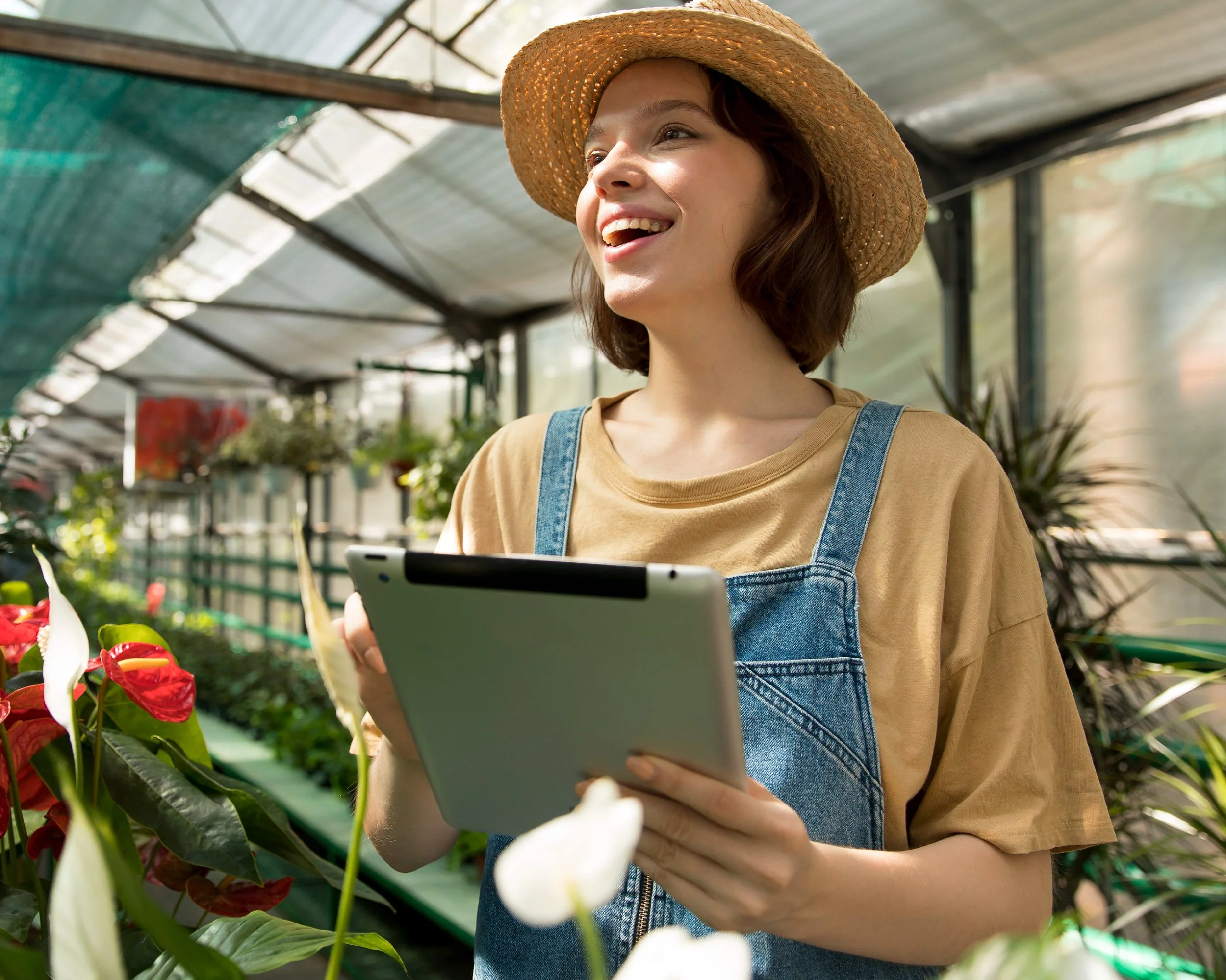 A young woman wearing a straw hat, beige t-shirt, and denim overalls stands in a greenhouse holding a tablet, smiling while tending to plants and flowers.
