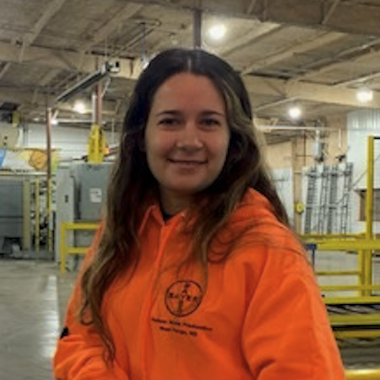 A young woman with long brown hair smiling, wearing an orange jacket with a logo, in an industrial or warehouse setting.