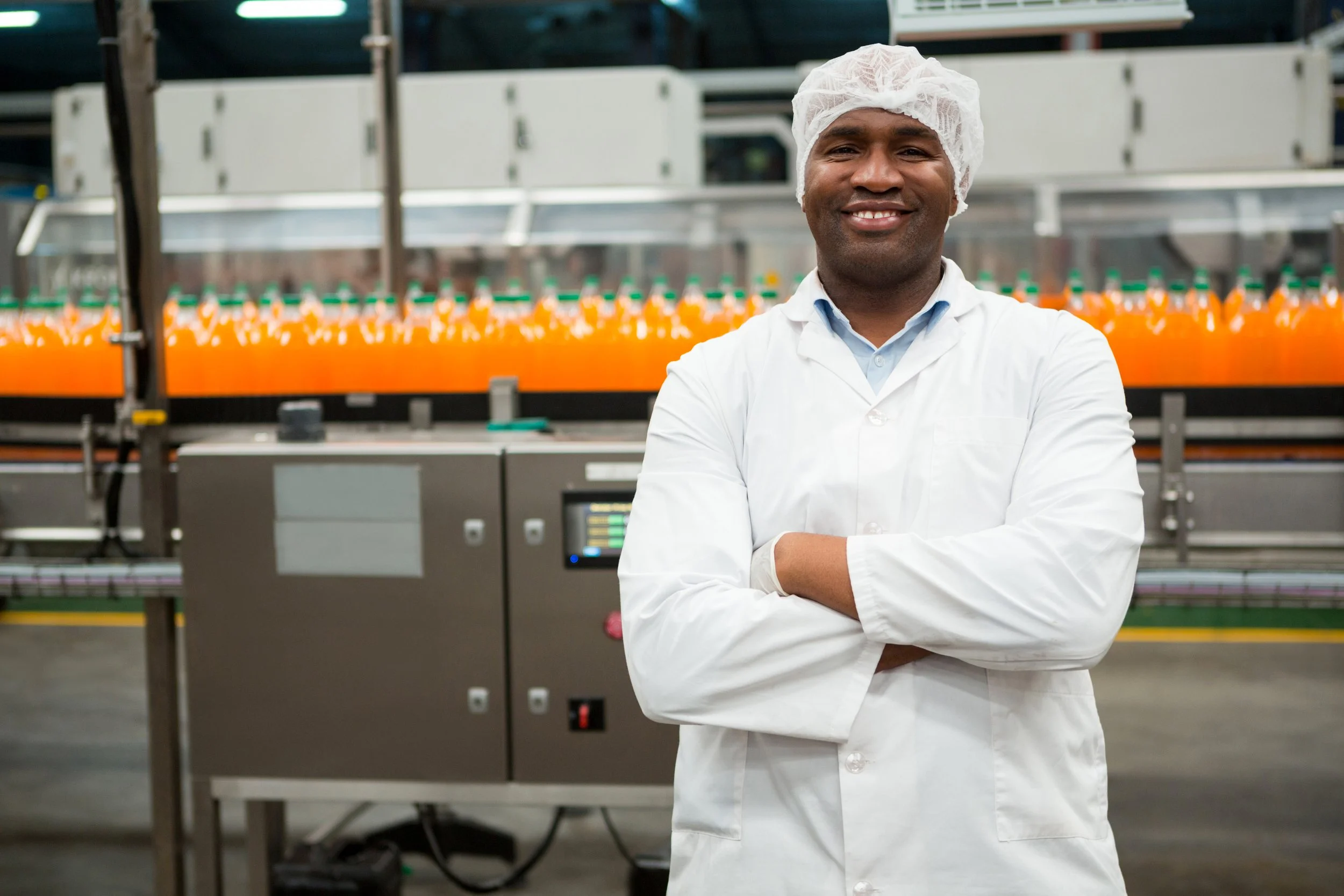 A smiling man in a white lab coat and hairnet stands with arms crossed in a beverage manufacturing facility, with bottles of orange soda on a conveyor belt in the background.