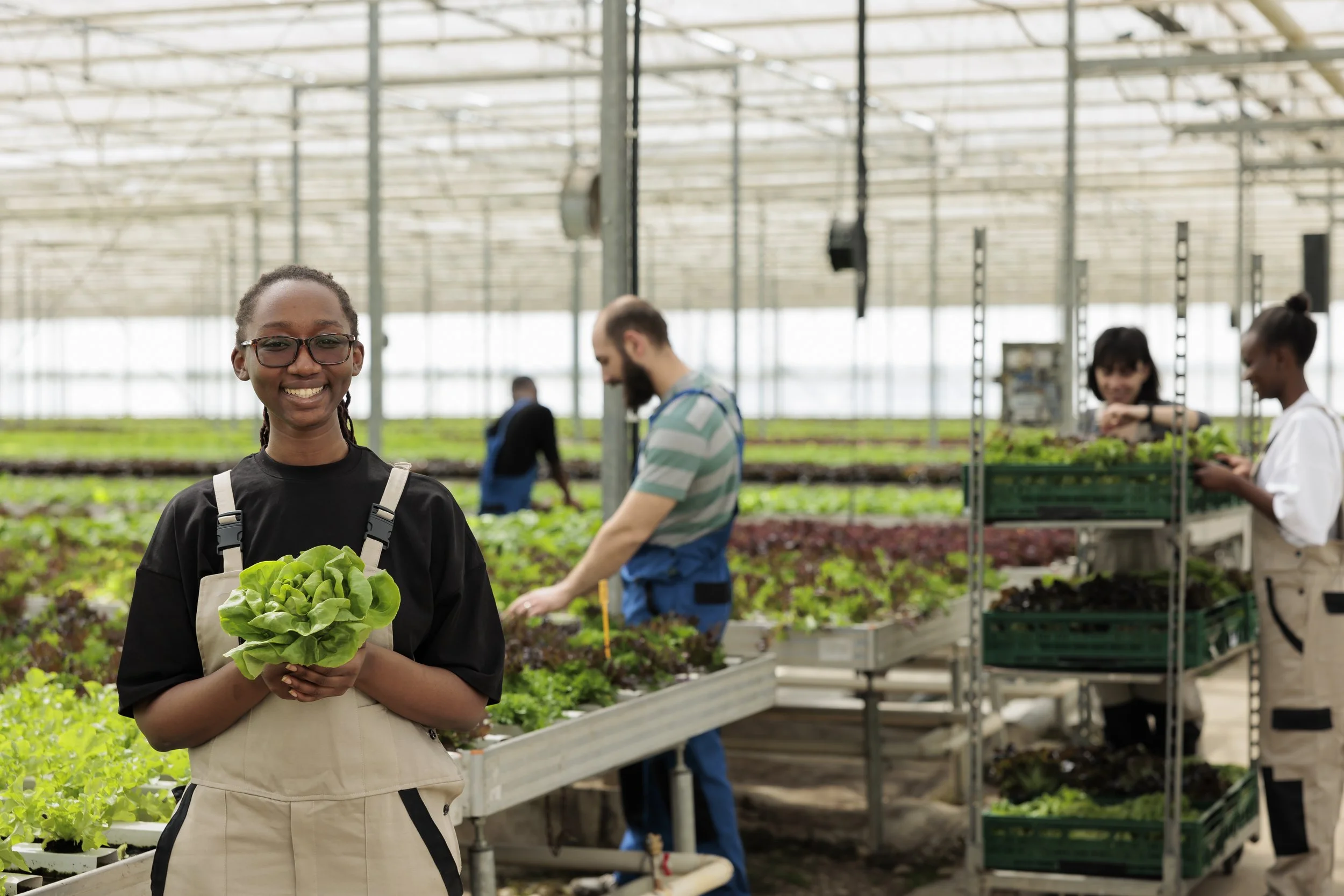 Smiling woman holding fresh lettuce in a greenhouse with other people working with plants in the background.