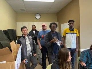 Five young men standing in a hallway, some holding papers. Several seated people are visible, with one woman in the foreground. The hallway has yellow walls, a clock on the wall, and boxes stacked on the left side.
