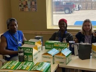 Three women sitting at a table with UN vaccine boxes and materials, in a room with a window showing a parking lot outside.