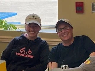 Two women sitting together indoors, both smiling, wearing casual clothes and hats. One is in a black hoodie with pink text, the other in a black T-shirt and glasses.