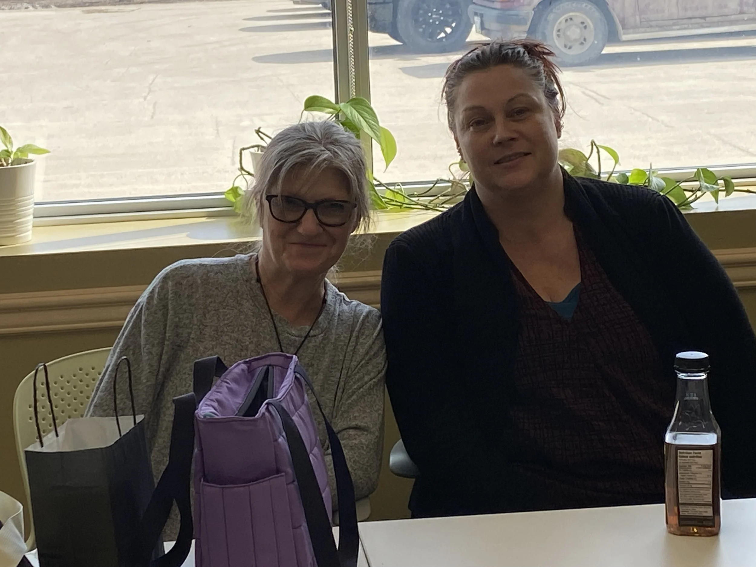 Two women sitting at a table near a window, with plants on the windowsill. One woman has short blonde hair, glasses, and a gray shirt. The other woman has red hair tied back, and is wearing a dark jacket over a shirt. On the table in front of them are a purple backpack, a gift bag, and a bottled drink.