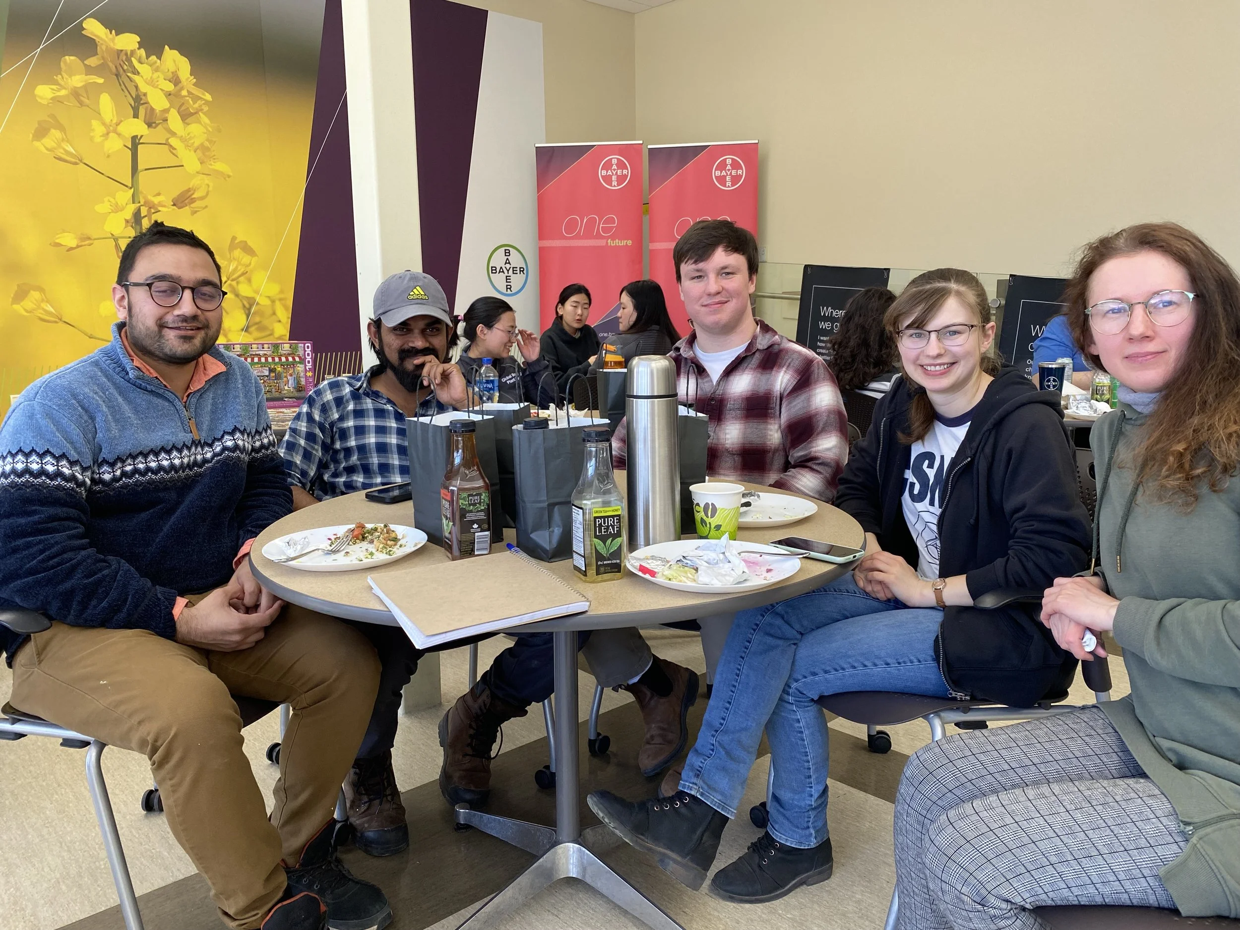 Group of six people sitting around a table during a meal in a cafeteria or conference setting, with half-eaten plates, drinks, and shopping bags on the table. Bright wall artwork and banners are in the background.