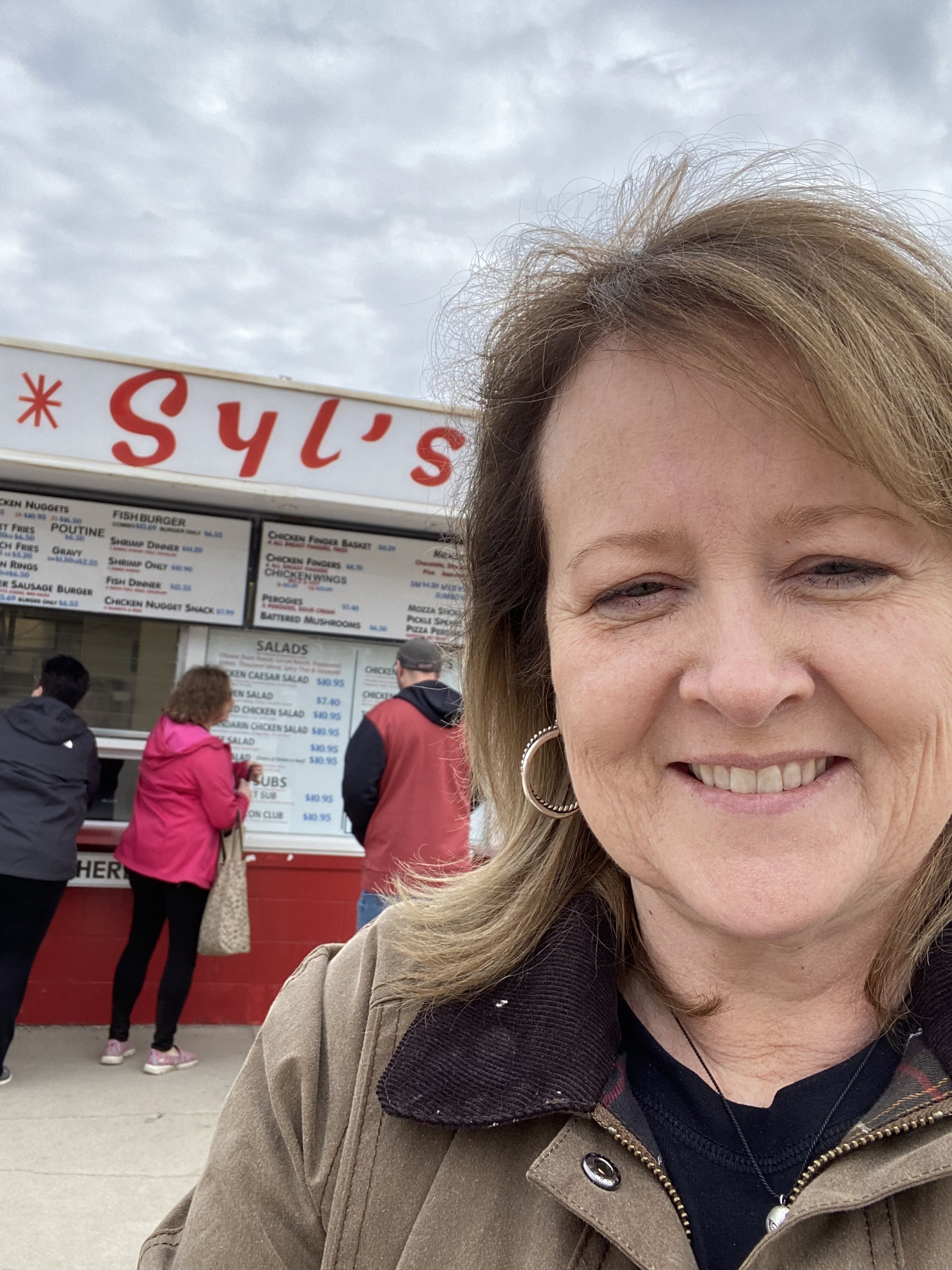 A woman smiling in front of a food stand with a menu, with customers ordering food, and cloudy sky overhead.