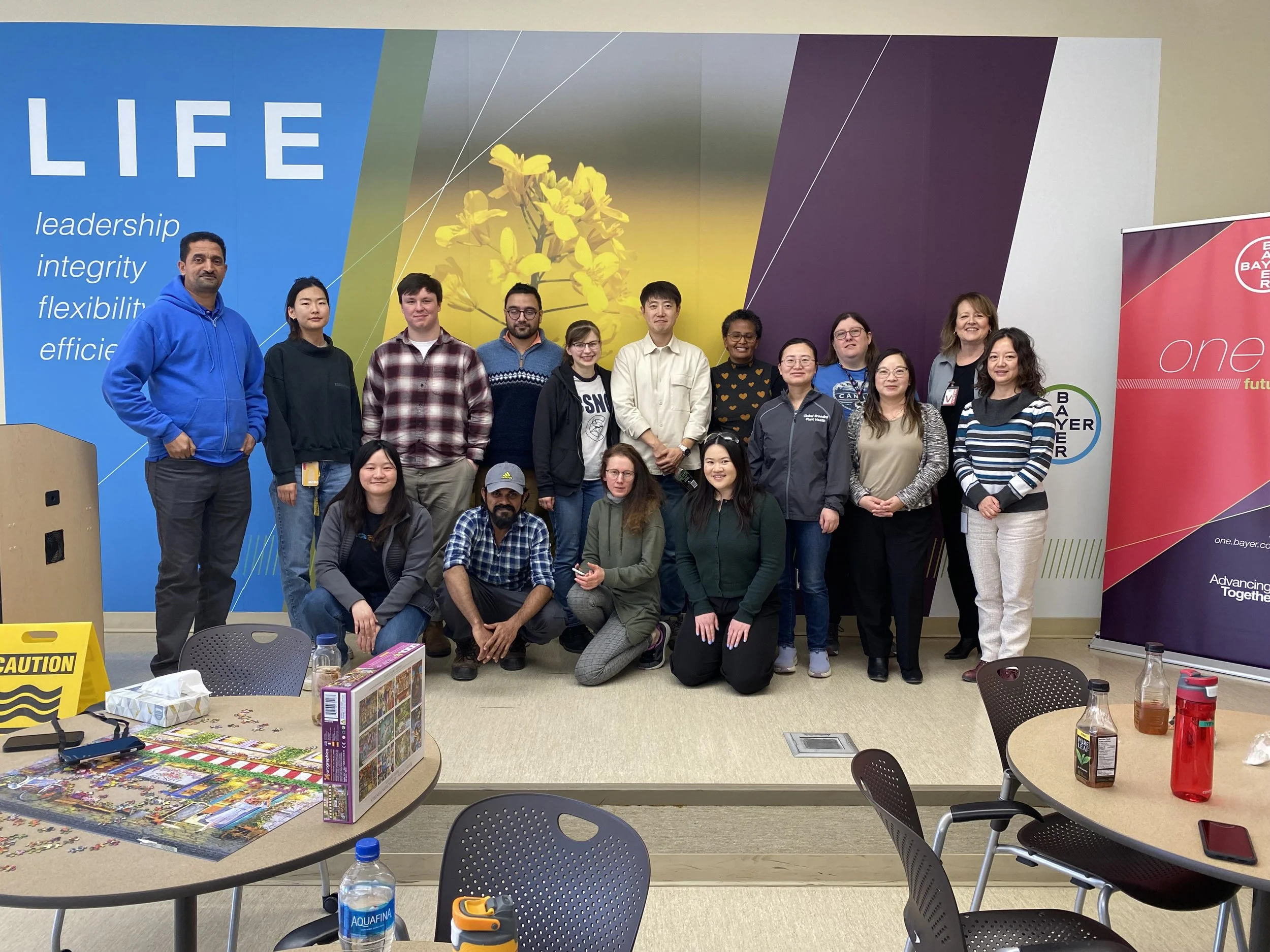 Group of 15 people posing for a photo in a room with colorful wall art and banners, tables with drinks and puzzle pieces on them.