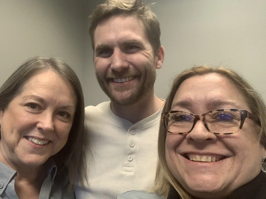 Three people smiling and taking a selfie indoors against a plain wall.